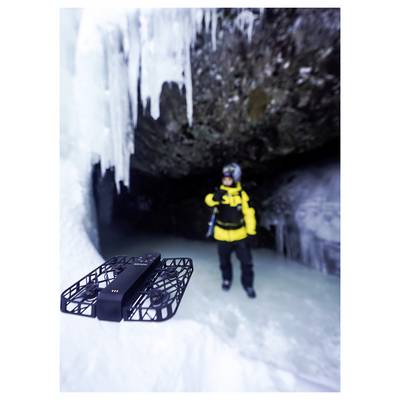 A person in winter gear stands in an icy cave, with icicles hanging from the ceiling, while a drone hovers in the foreground.