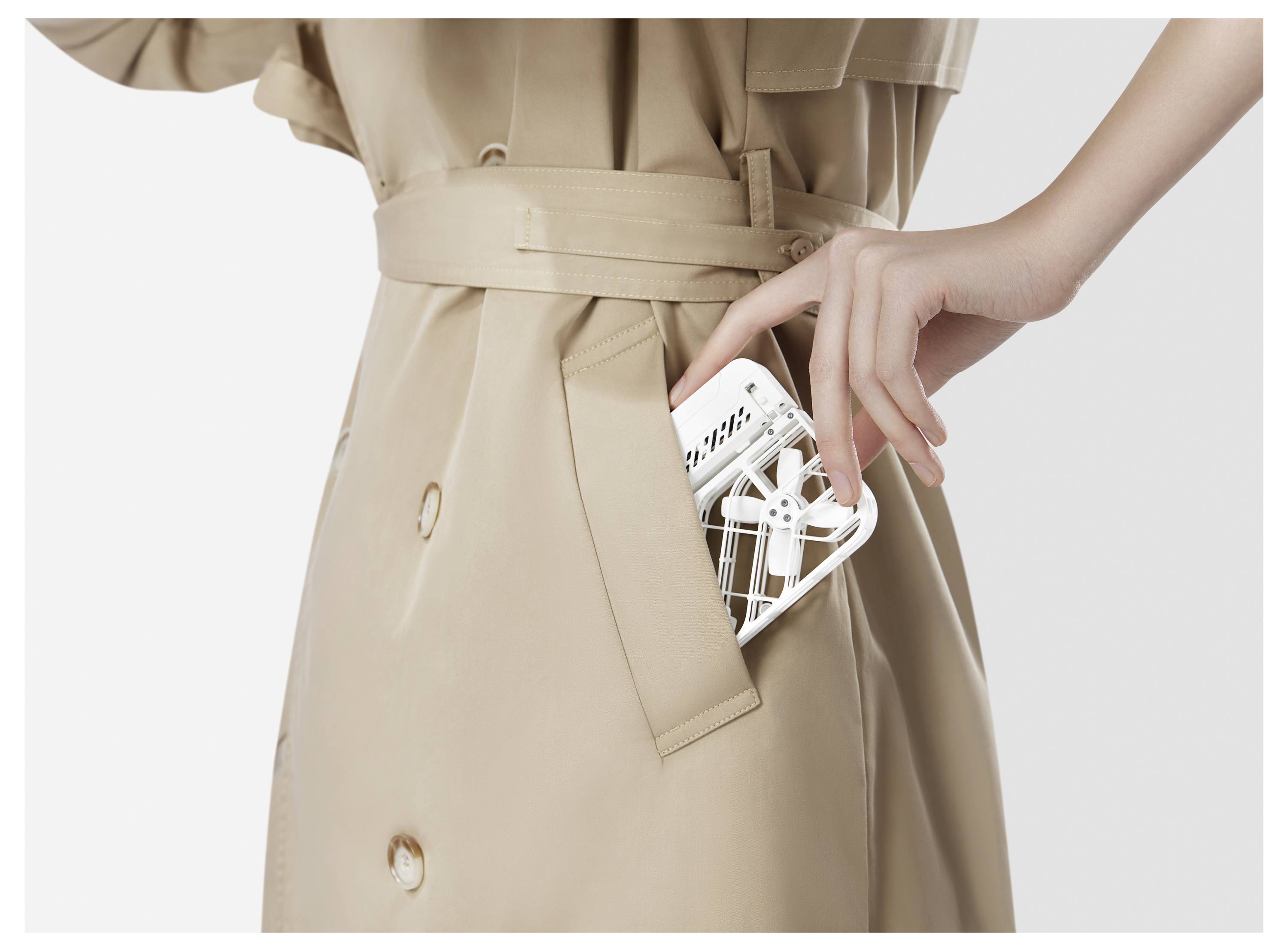 A person in a beige trench coat places a small white drone into their coat pocket.