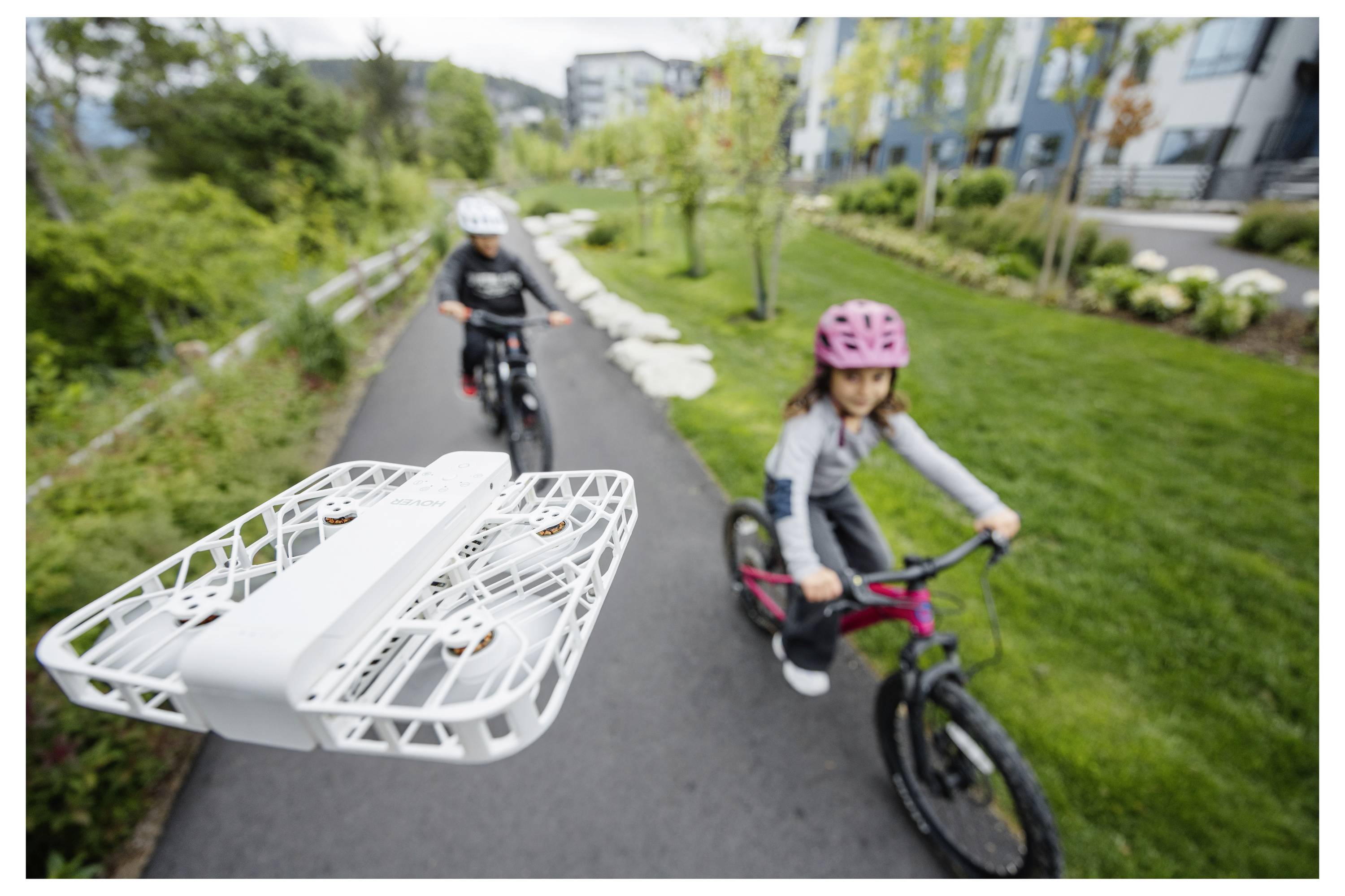 A camera drone flies above a pathway capturing footage of two children riding bicycles in a park setting.