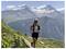 A person is running on a mountain trail surrounded by greenery, with snow-capped peaks in the background under a clear blue sky.