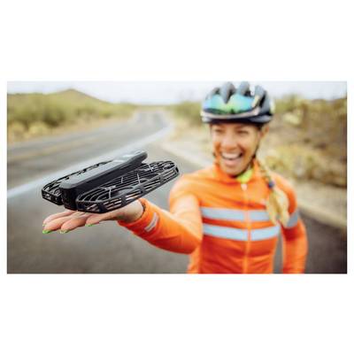 A cyclist in an orange jacket holds a small drone on an open road, smiling and wearing a helmet, with a blurred desert background.