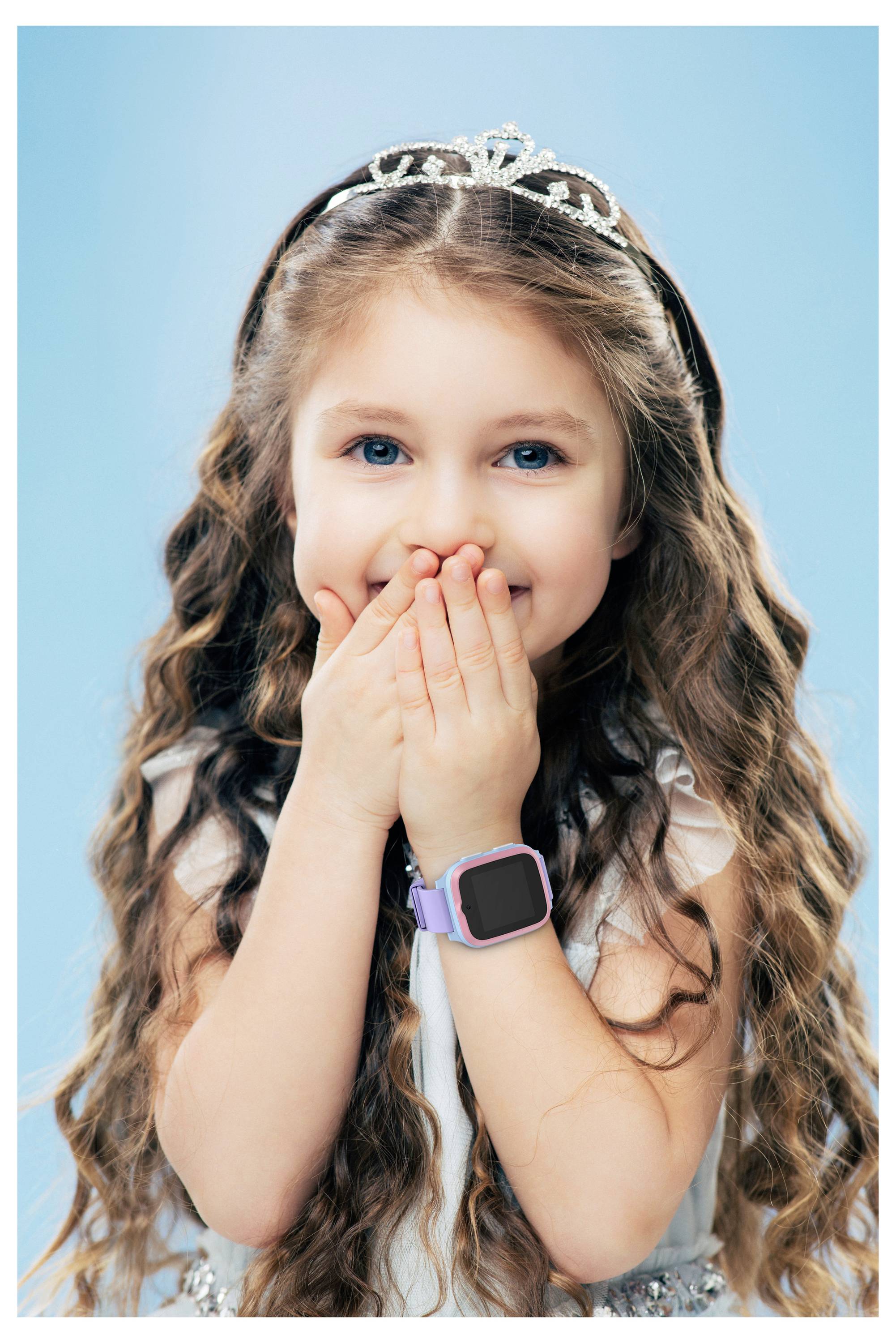 A young girl with long curly hair and a crown, covering her mouth with both hands, wearing a smartwatch, against a blue background.