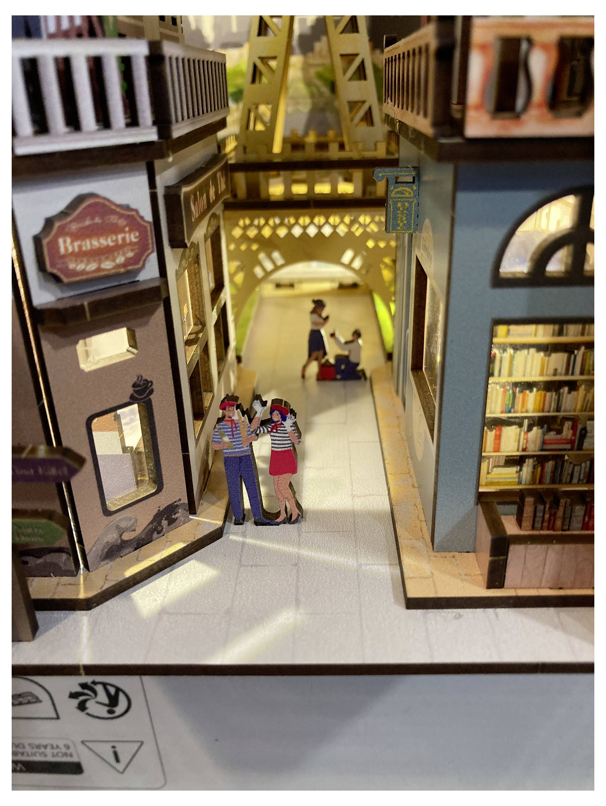 A miniature model depicting a Parisian street scene with a bookshop, brasserie, and the Eiffel Tower in the background. People are strolling.