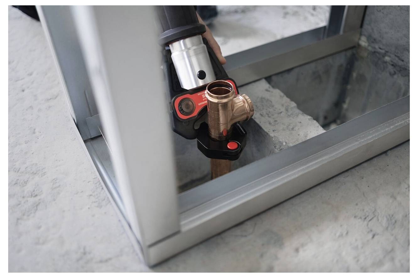 A close-up of a hand using a pipe pressing tool to connect copper piping within a metal frame on a concrete floor.