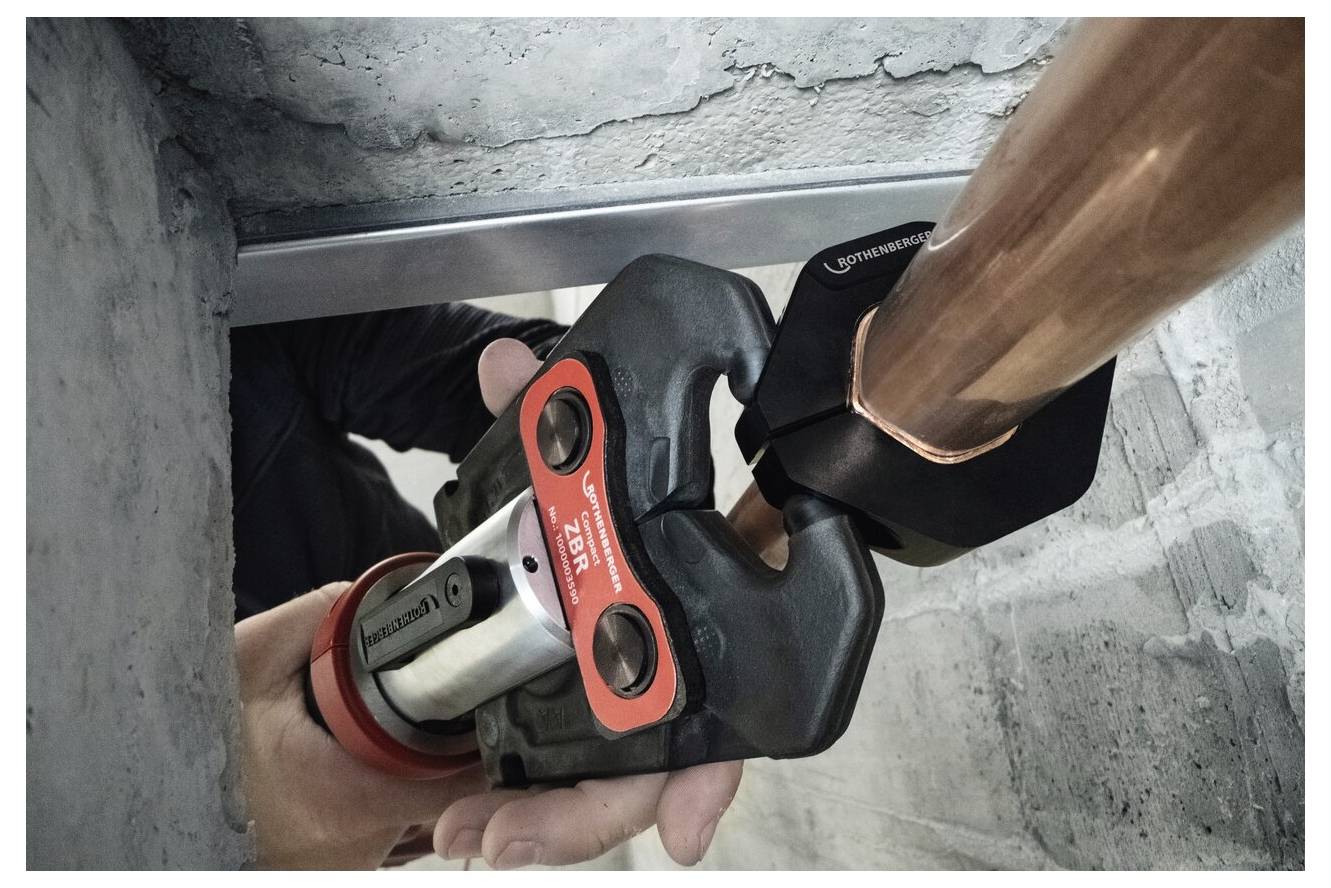 A person uses a hydraulic tool to join a copper pipe in a concrete wall. The tool grips tightly, ensuring a secure connection.