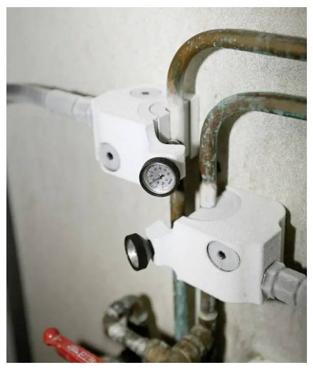 Close-up of two pipe shutoff valves on copper plumbing, with a pressure gauge and red handle visible against a wall.