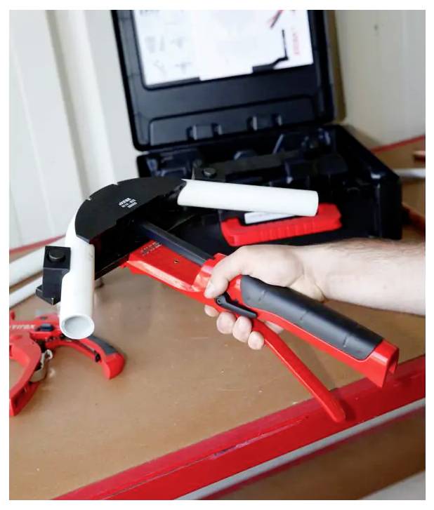 Person holding a red-and-black pipe cutter over a workbench, with additional plumbing tools nearby in an open case.