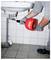 Plumber using a red Virax drain auger to unclog a bathroom sink drain beneath a wall-mounted basin.<br>
