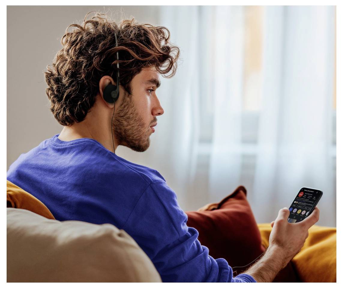 A person wearing headphones is sitting on a couch and looking at a smartphone screen in a cozy, well-lit room.
