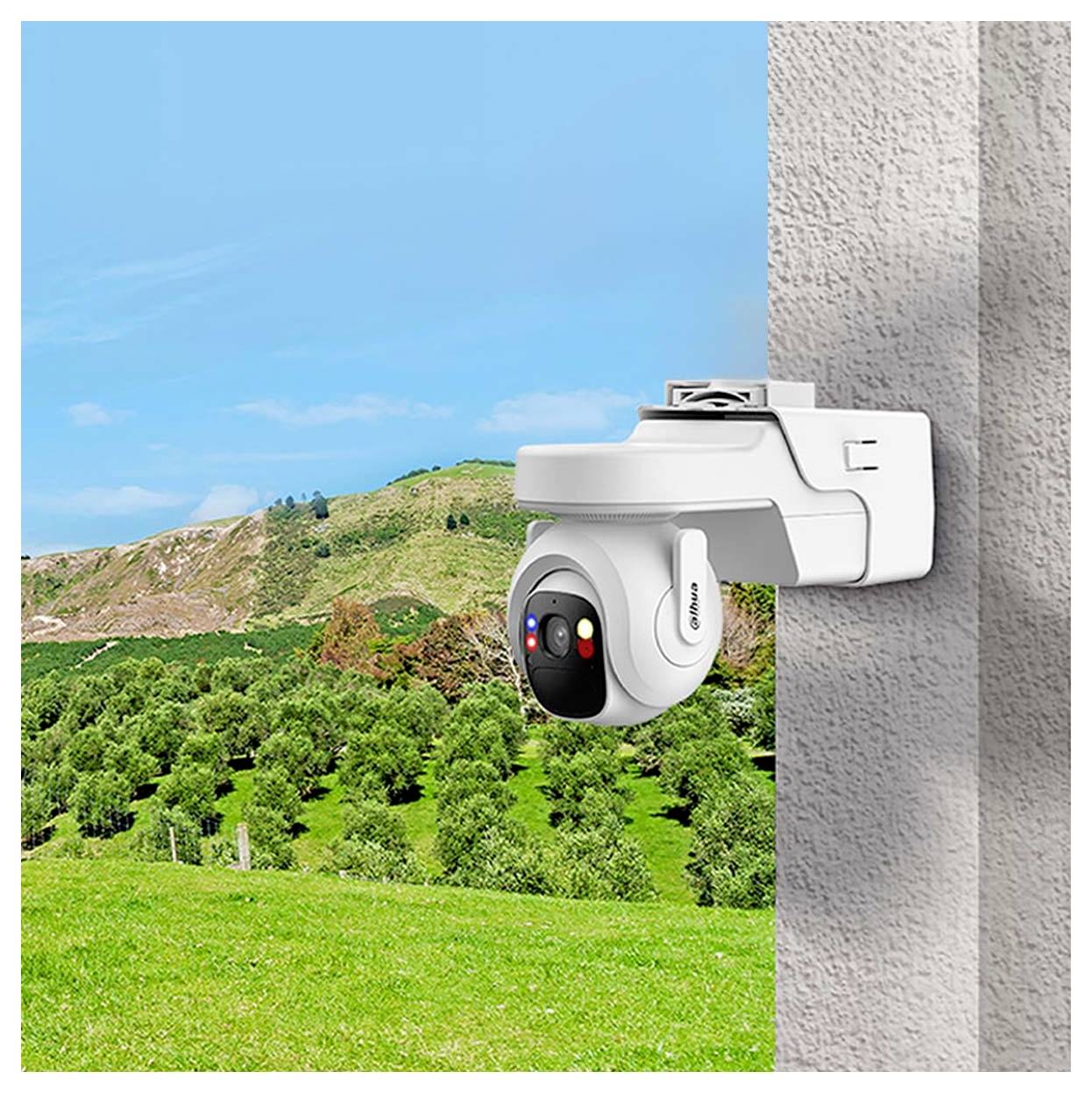 A white security camera is mounted on a building wall overlooking a green landscape with hills and a blue sky in the background.