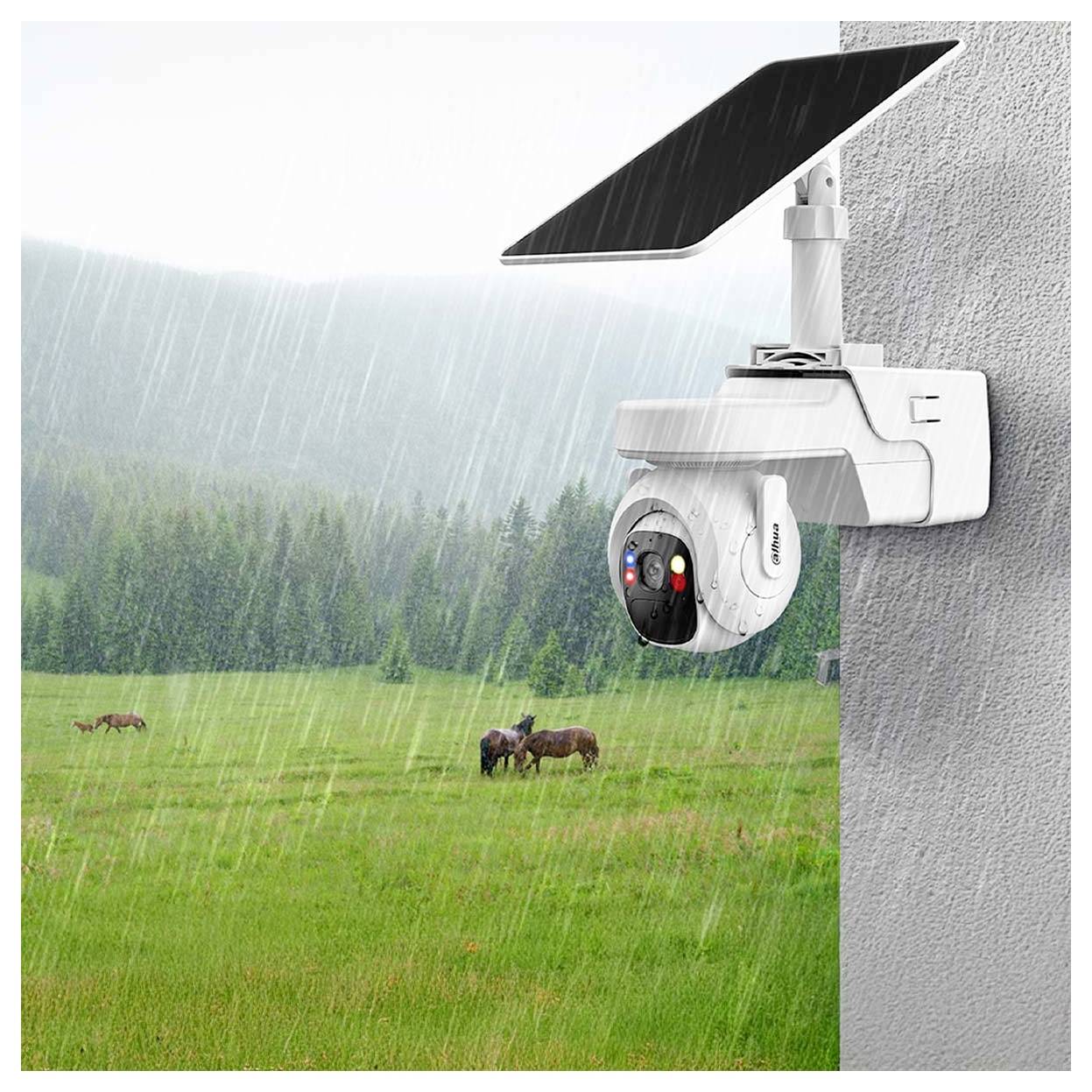 A solar-powered security camera mounted on a wall overlooks a rainy, grassy field with grazing deer in the background.