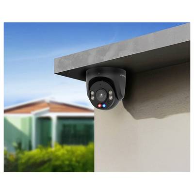 A black security camera is mounted on an outdoor wall, overlooking a suburban area with blurred houses and greenery in the background.