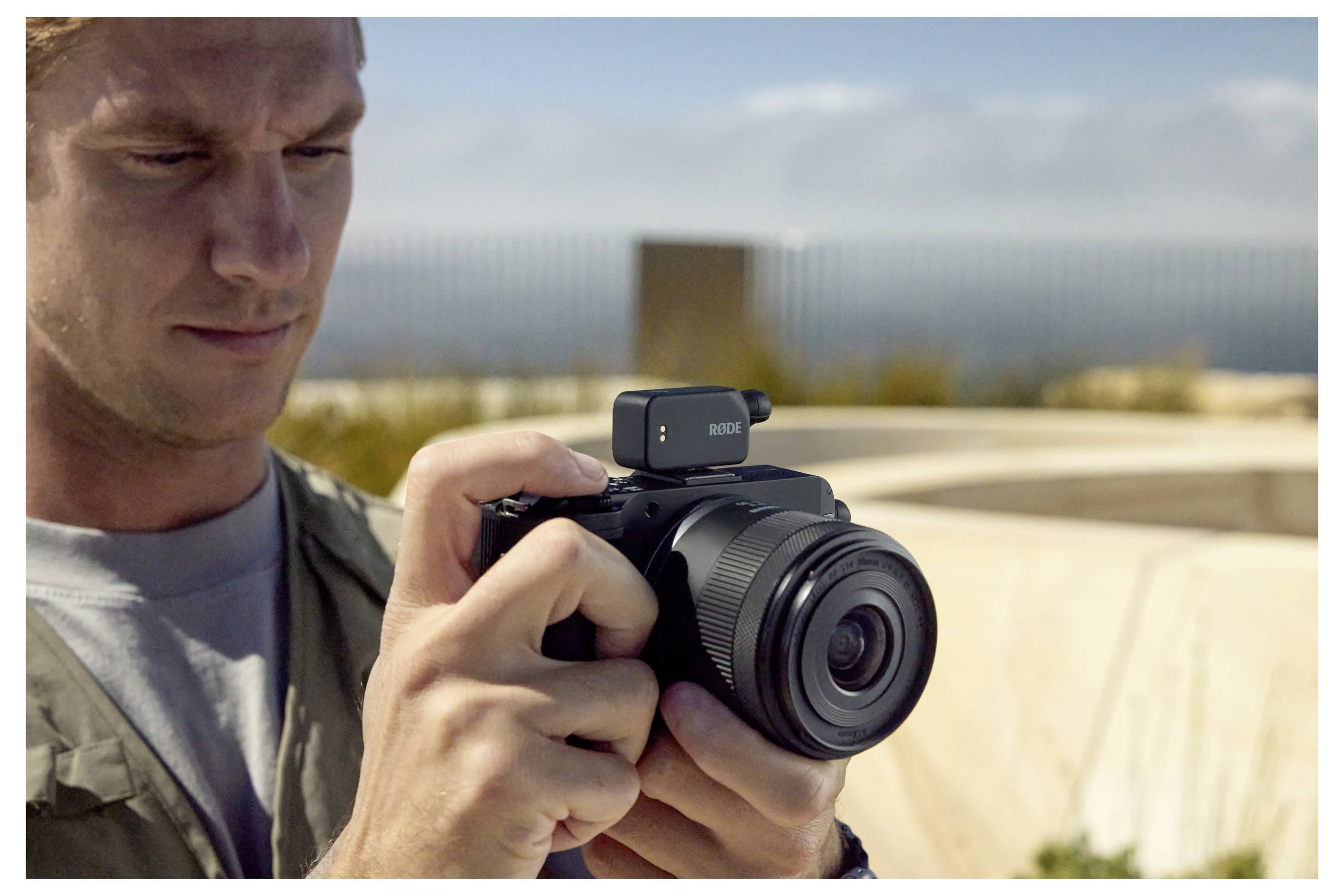 A person outdoors focuses intently on adjusting a camera with a small microphone attached, set against a blurred backdrop of sky and structures.
