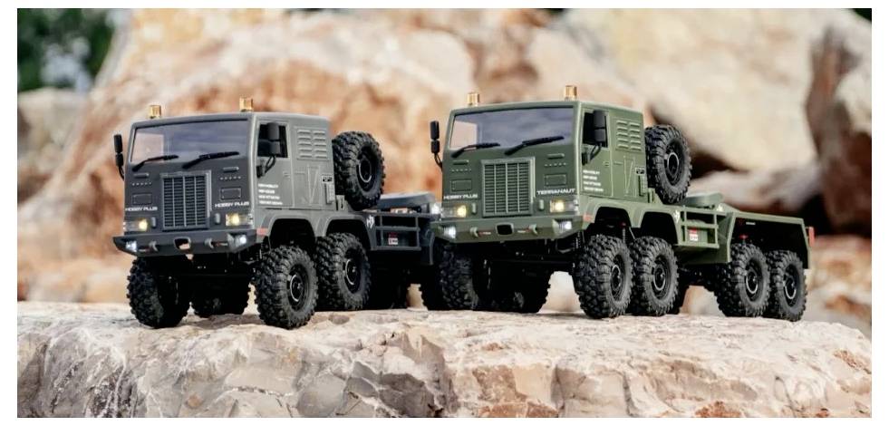 Two model off-road trucks, one black and one green, with six wheels each, are displayed on a rocky terrain with a blurred background.