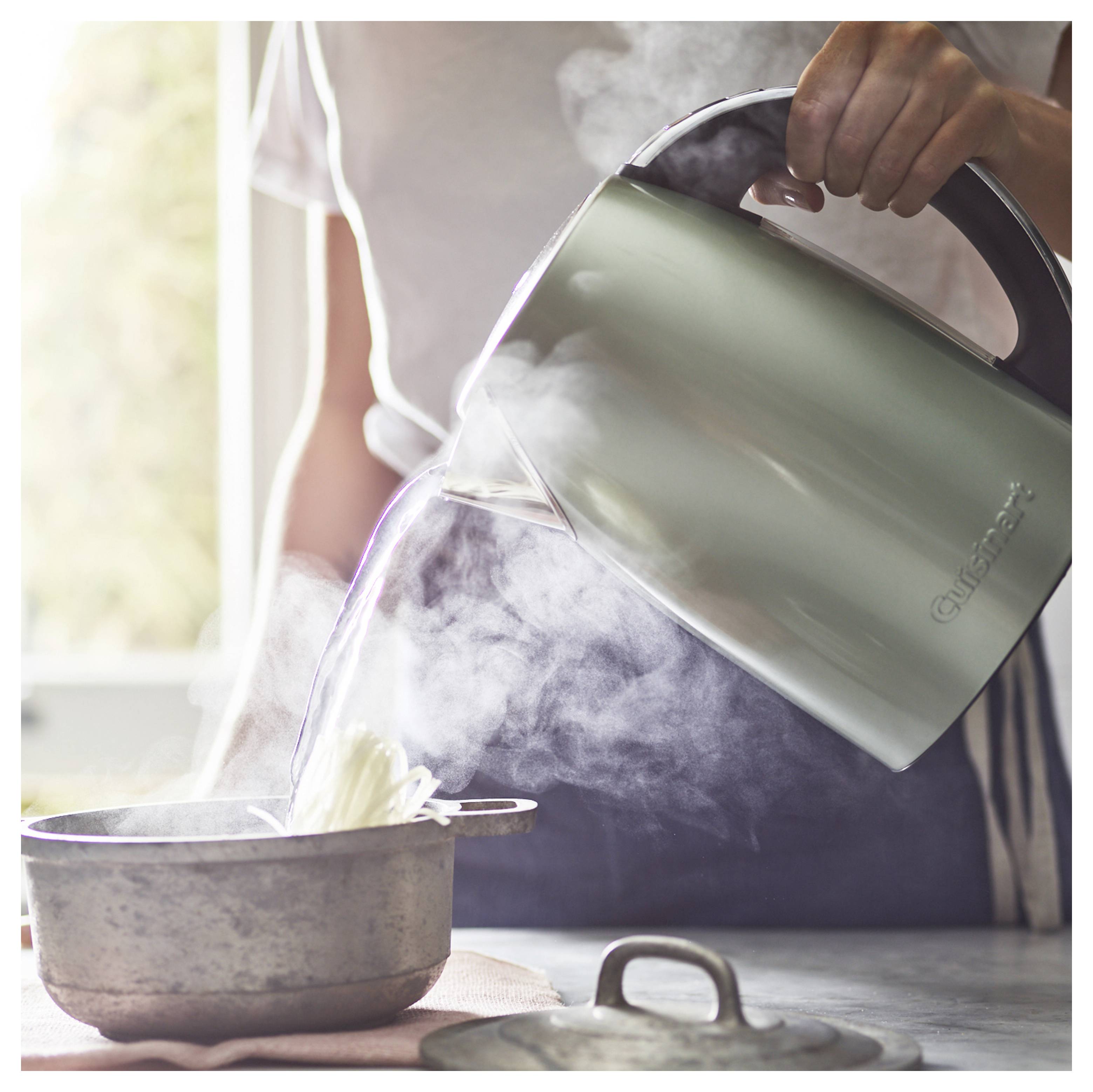 A person pours hot water from a kettle into a pot with steam rising, suggesting cooking or food preparation in a kitchen setting.