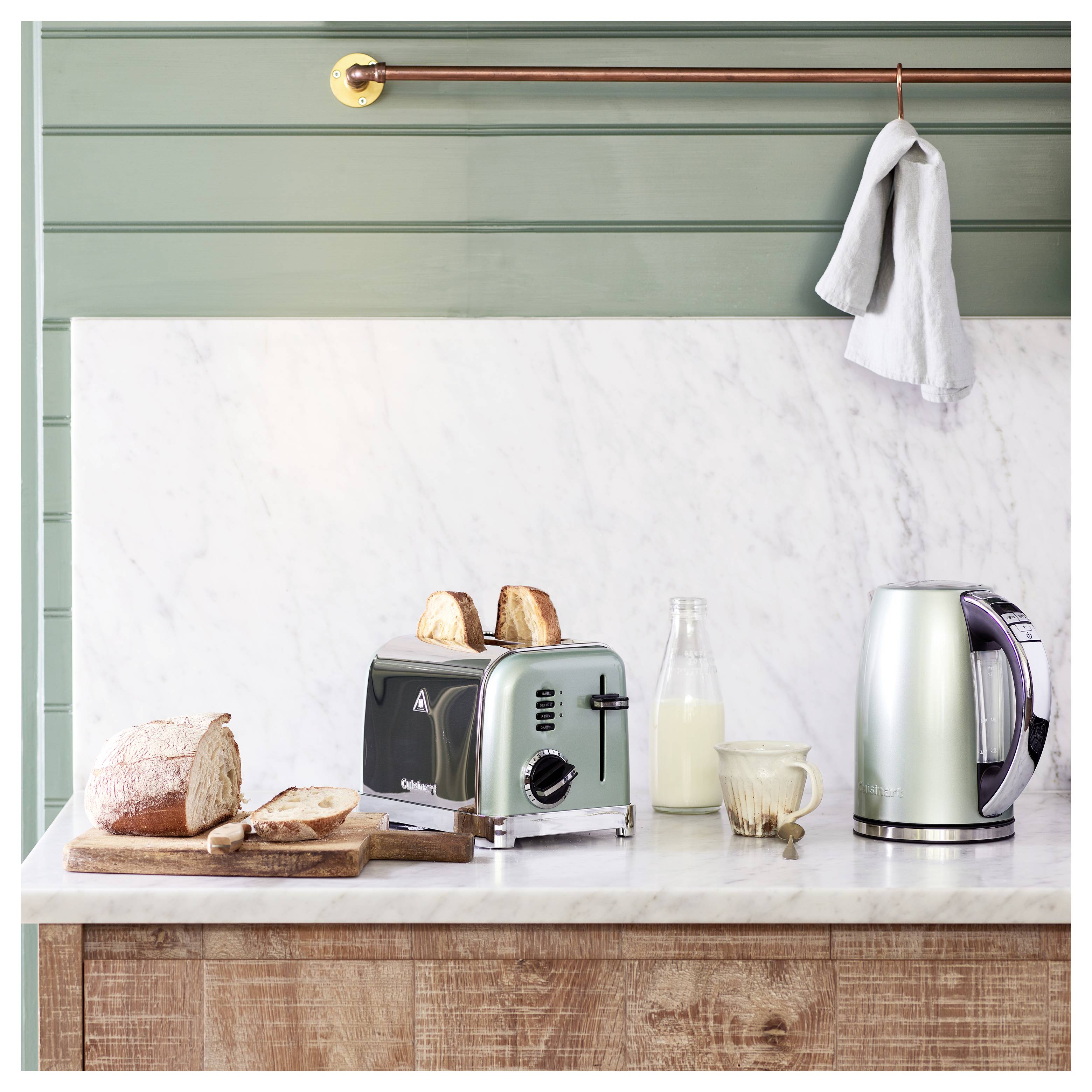 A kitchen counter with a loaf of sliced bread, a toaster with toast, a bottle and glass of milk, and an electric kettle against a green wall.