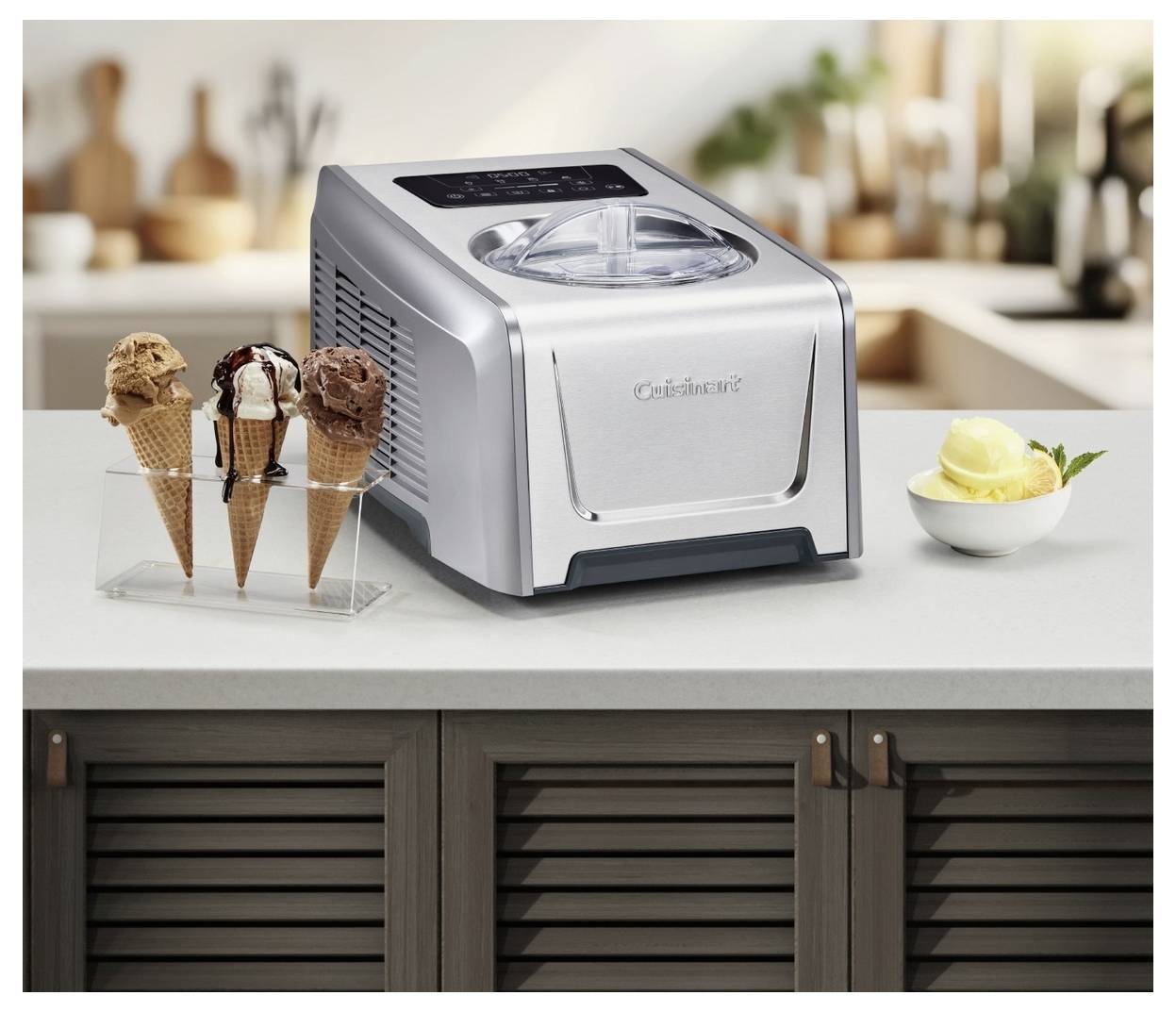 A countertop ice cream maker sits on a kitchen surface beside a dish of yellow ice cream and three ice cream cones in a holder.