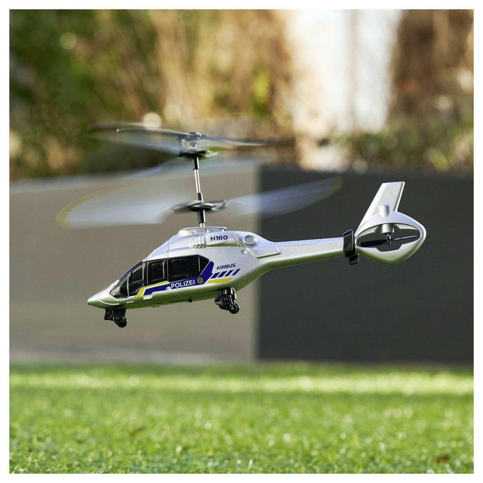 A remote-controlled helicopter with 'Polizei' and 'H160' markings hovers low above grassy ground, with blurry trees in the background.