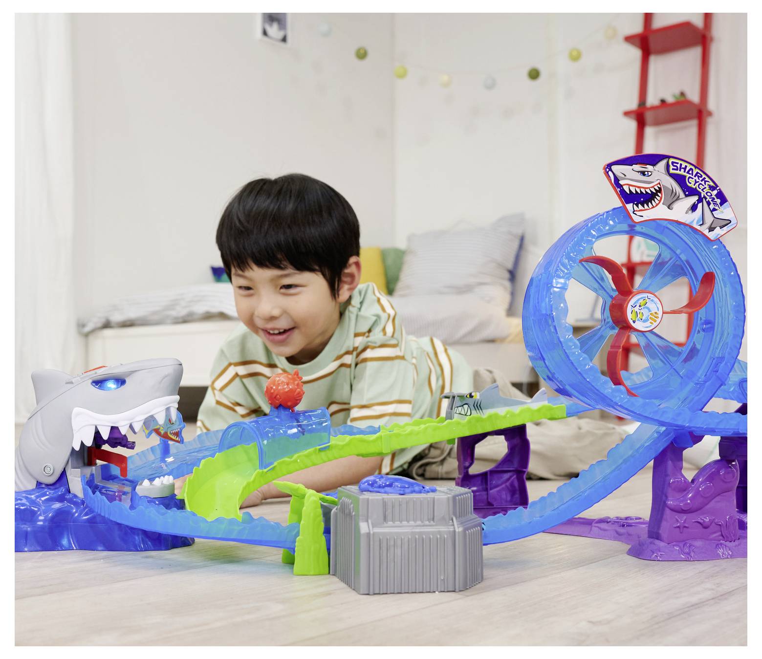 A child plays with a toy racetrack featuring a loop and a shark head. The child watches a small car navigating the track.