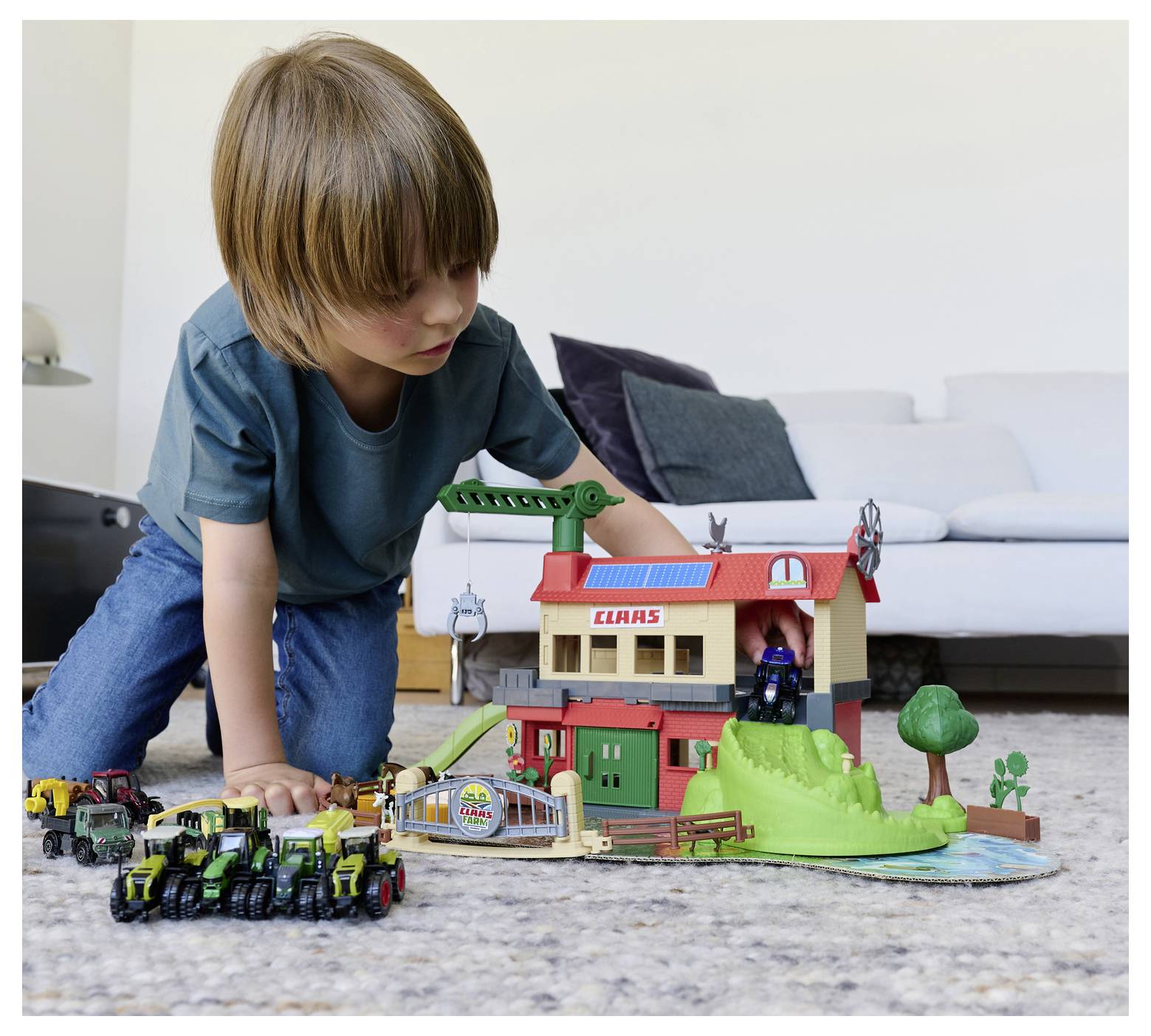 A child plays with a toy farm set, featuring a barn labeled 'CLAAS' and several toy tractors on a carpeted floor, near a sofa.