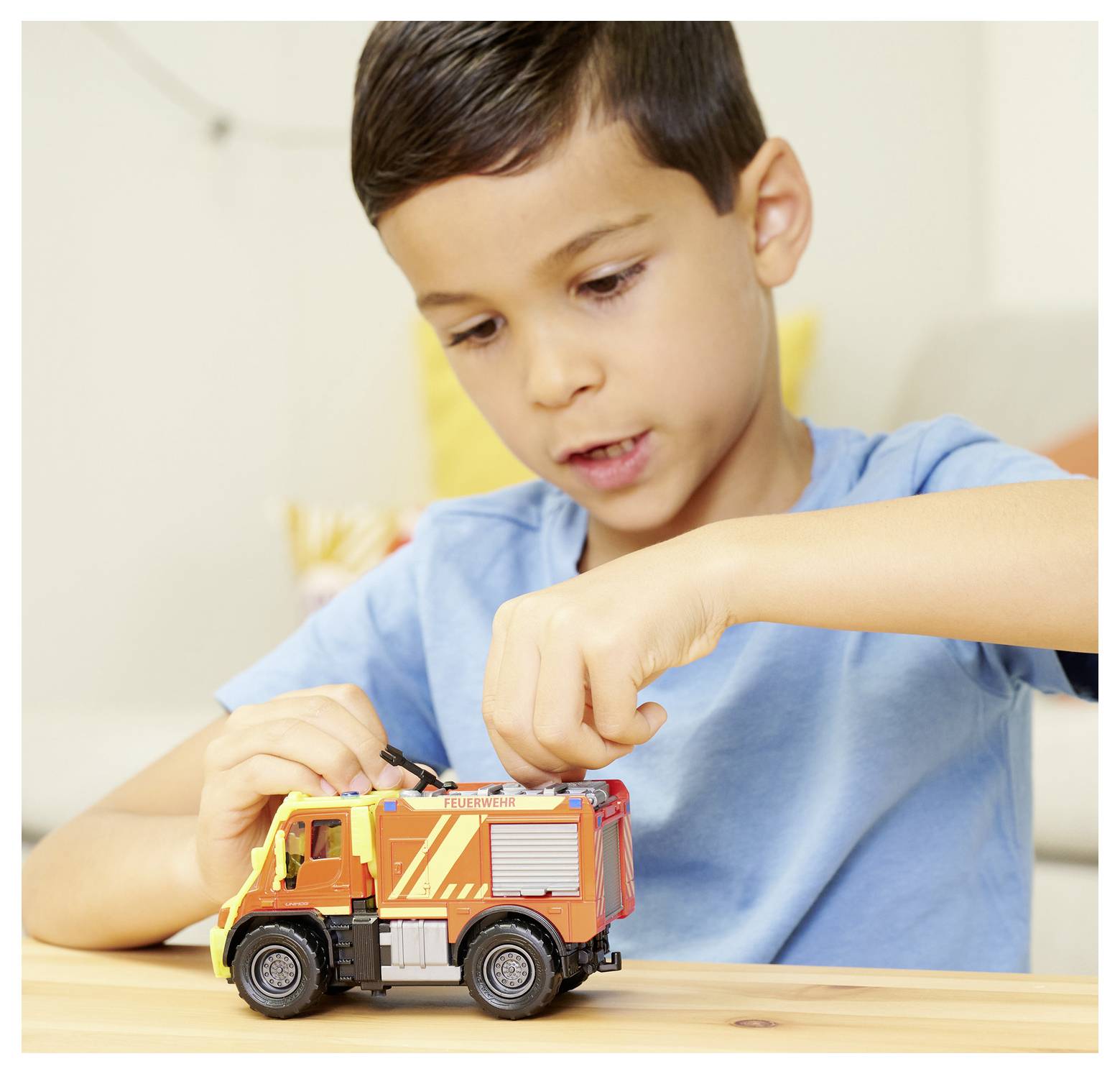 A young boy in a blue shirt plays with a toy fire truck at a table, focusing intently on the toy.