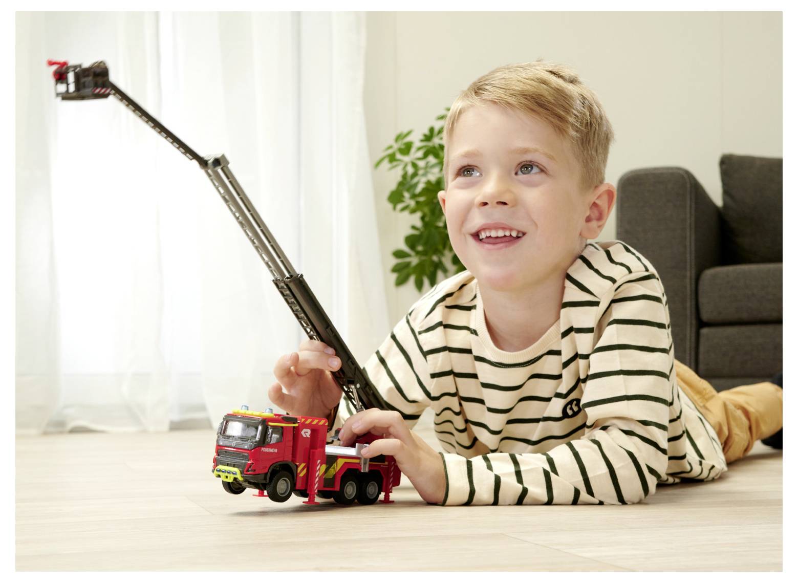 A young child, smiling and lying on the floor, plays with a toy fire truck with an extended ladder in a cozy living room setting.