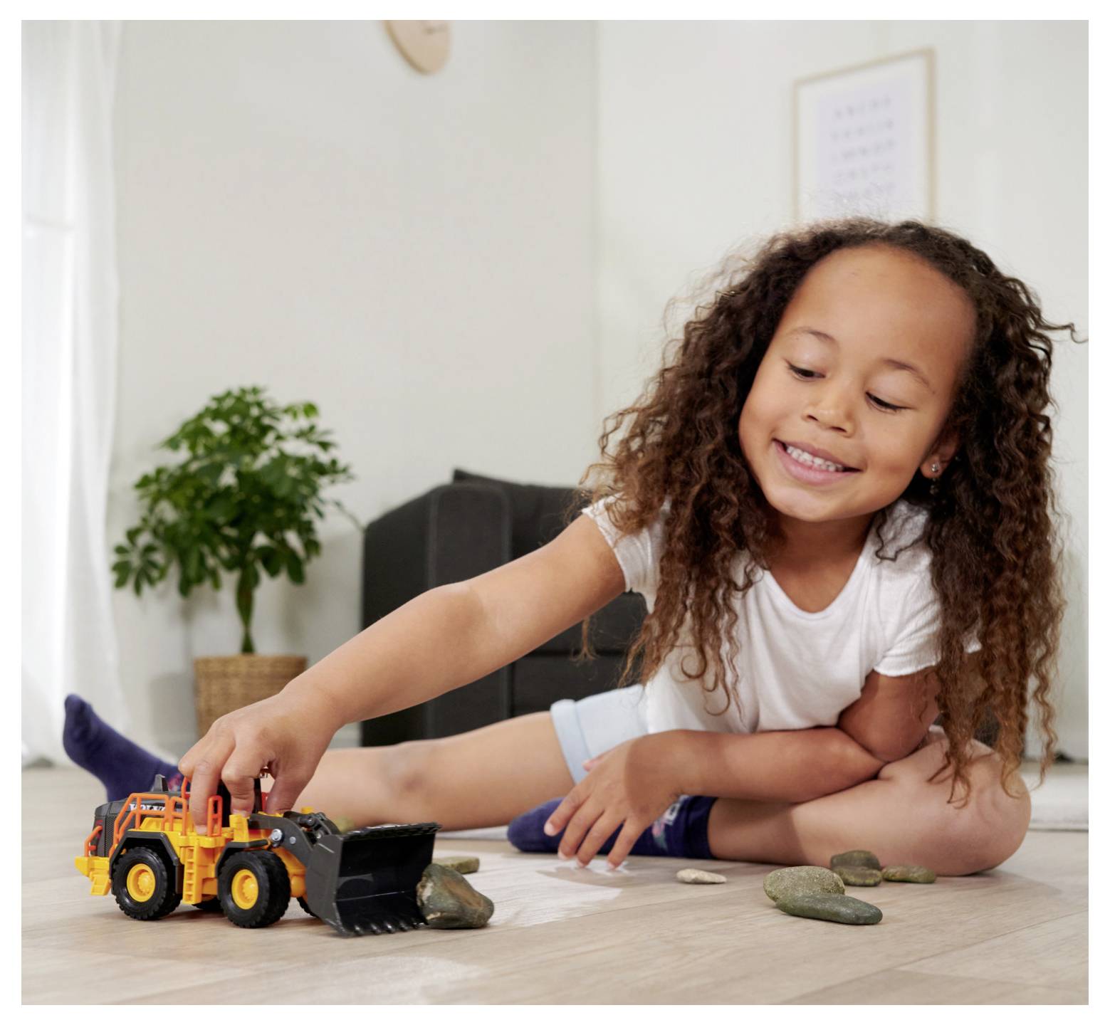 A child with curly hair is happily playing indoors with a yellow toy bulldozer, moving small rocks on a wooden floor.