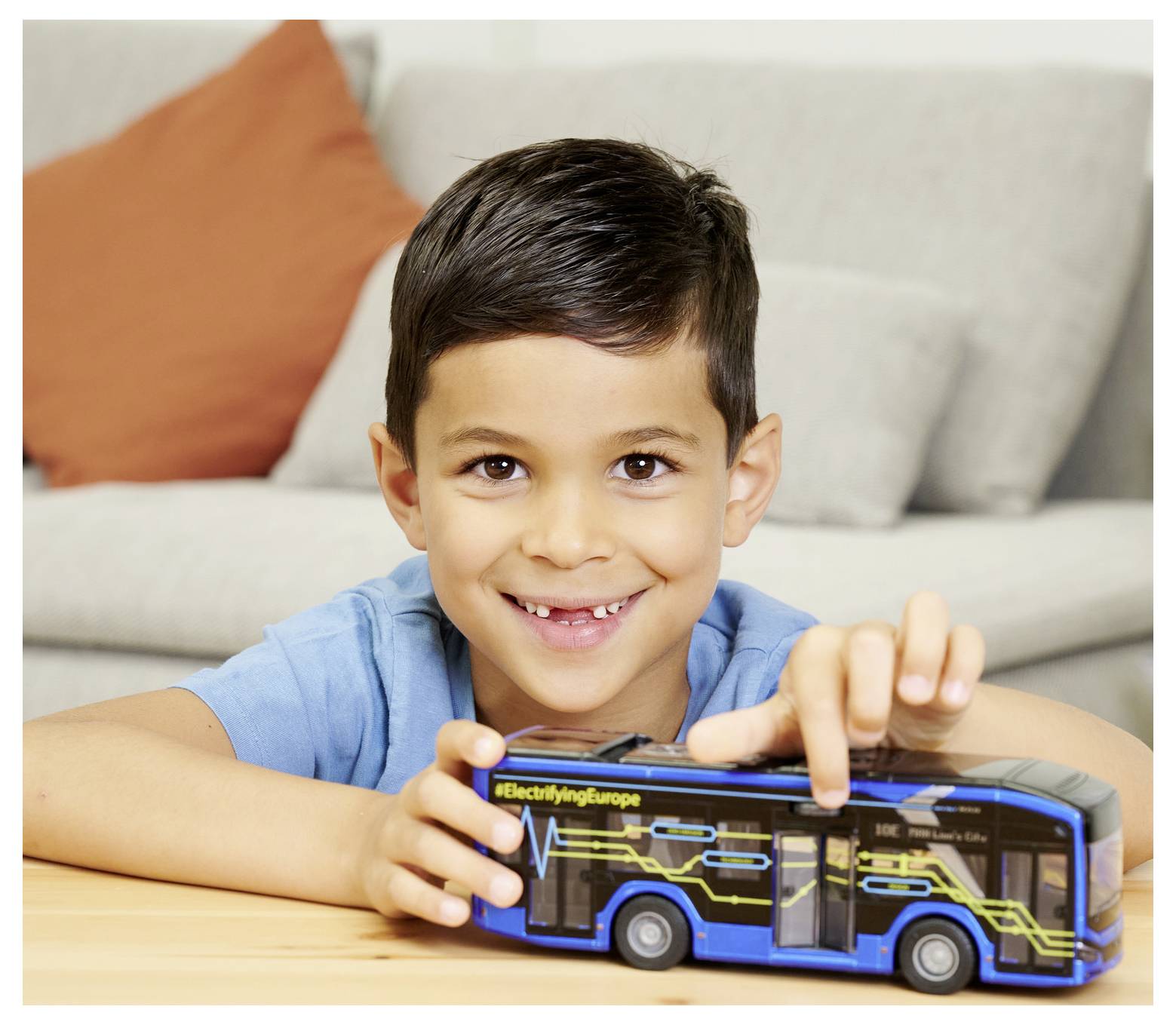 A child smirks while holding a toy bus with the text '#ElectrifyingEurope' on it, sitting in a living room setting.