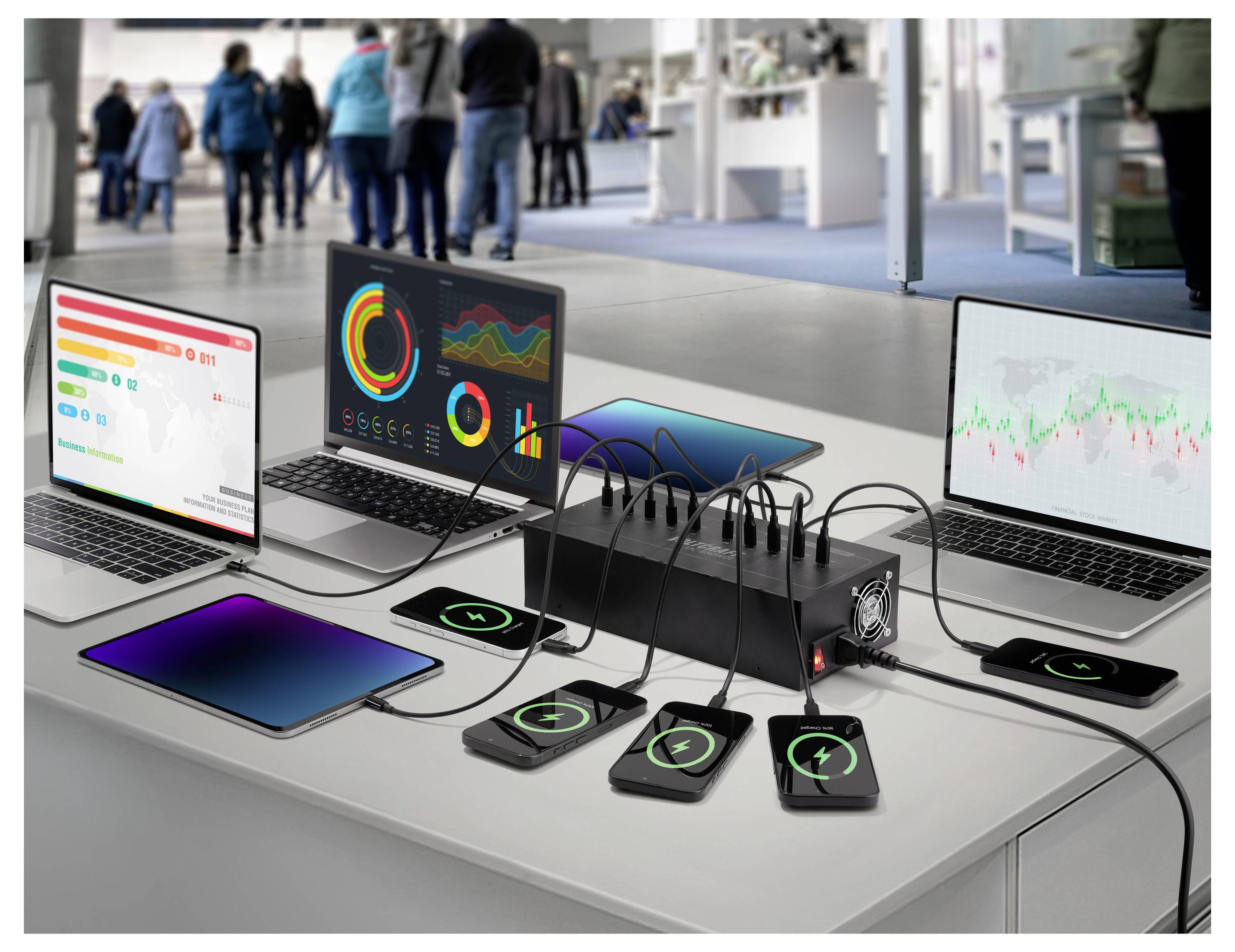 Three laptops display charts and graphs. Phones charge wirelessly on a table with a multi-device charger. Blurred figures in background.