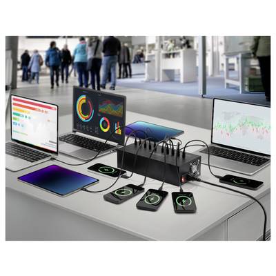 Three laptops display charts and graphs. Phones charge wirelessly on a table with a multi-device charger. Blurred figures in background.