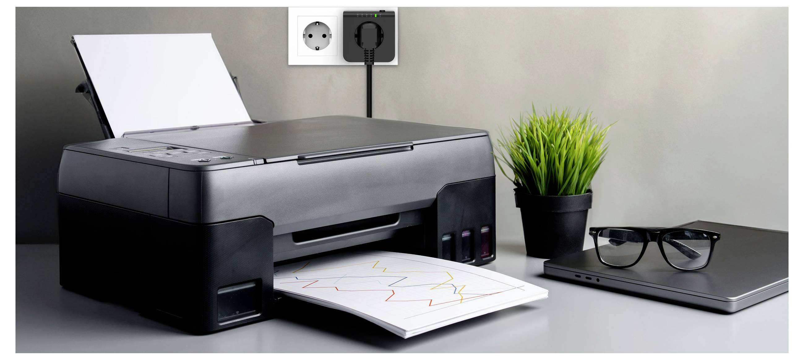 A printer on a desk with a printed page partly ejected, next to a laptop, glasses, and a small potted plant. An outlet is in the background.