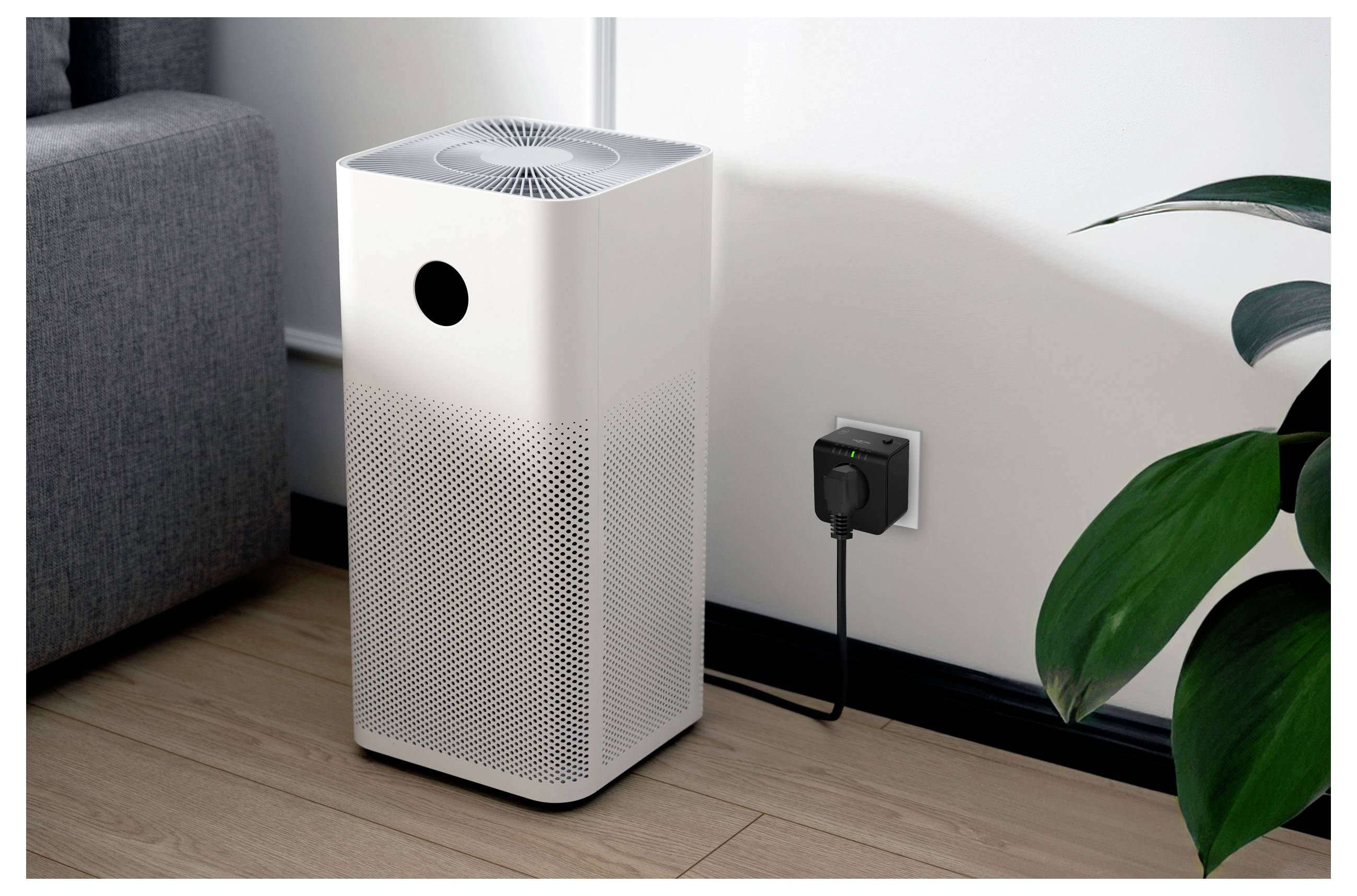 'White air purifier' placed on a wooden floor next to a gray sofa, plugged into a wall outlet, near a green plant.