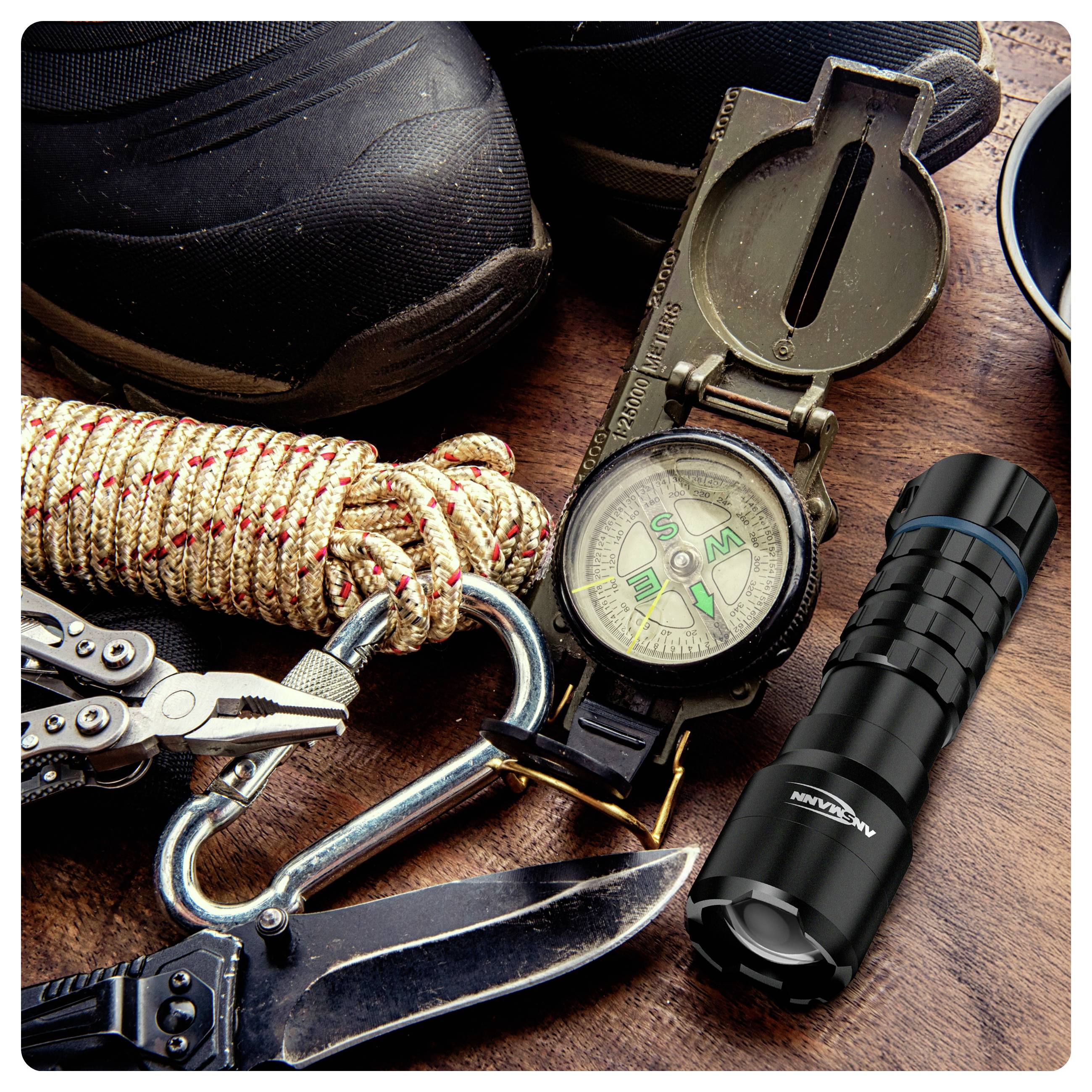 Outdoor gear including a hiking boot, compass, flashlight, multitool, carabiner, rope, and knife on a wooden surface, suggesting readiness for adventure.