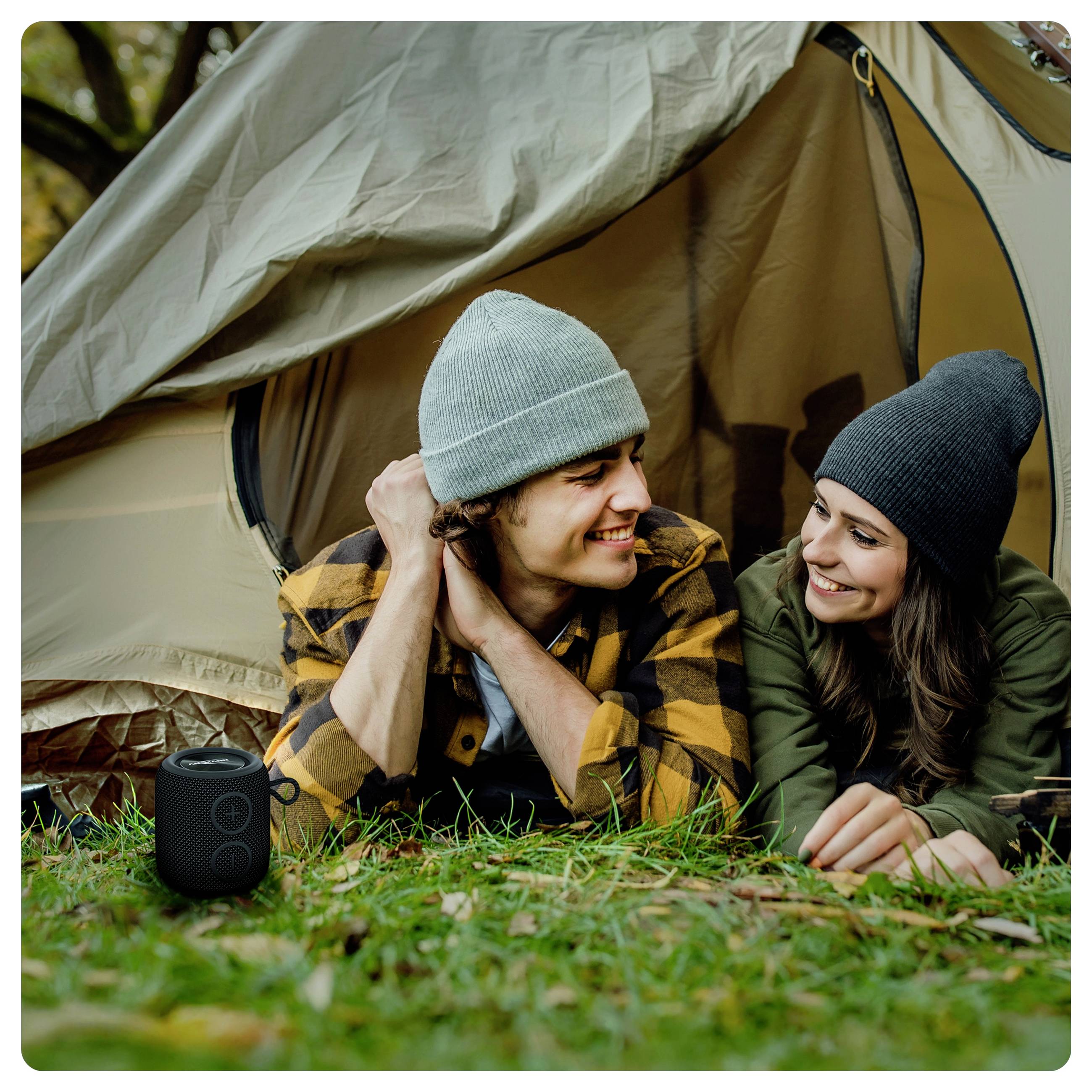A couple in beanies and warm clothing lies on grass, smiling at each other by an open tent in a forest. A black speaker is nearby.