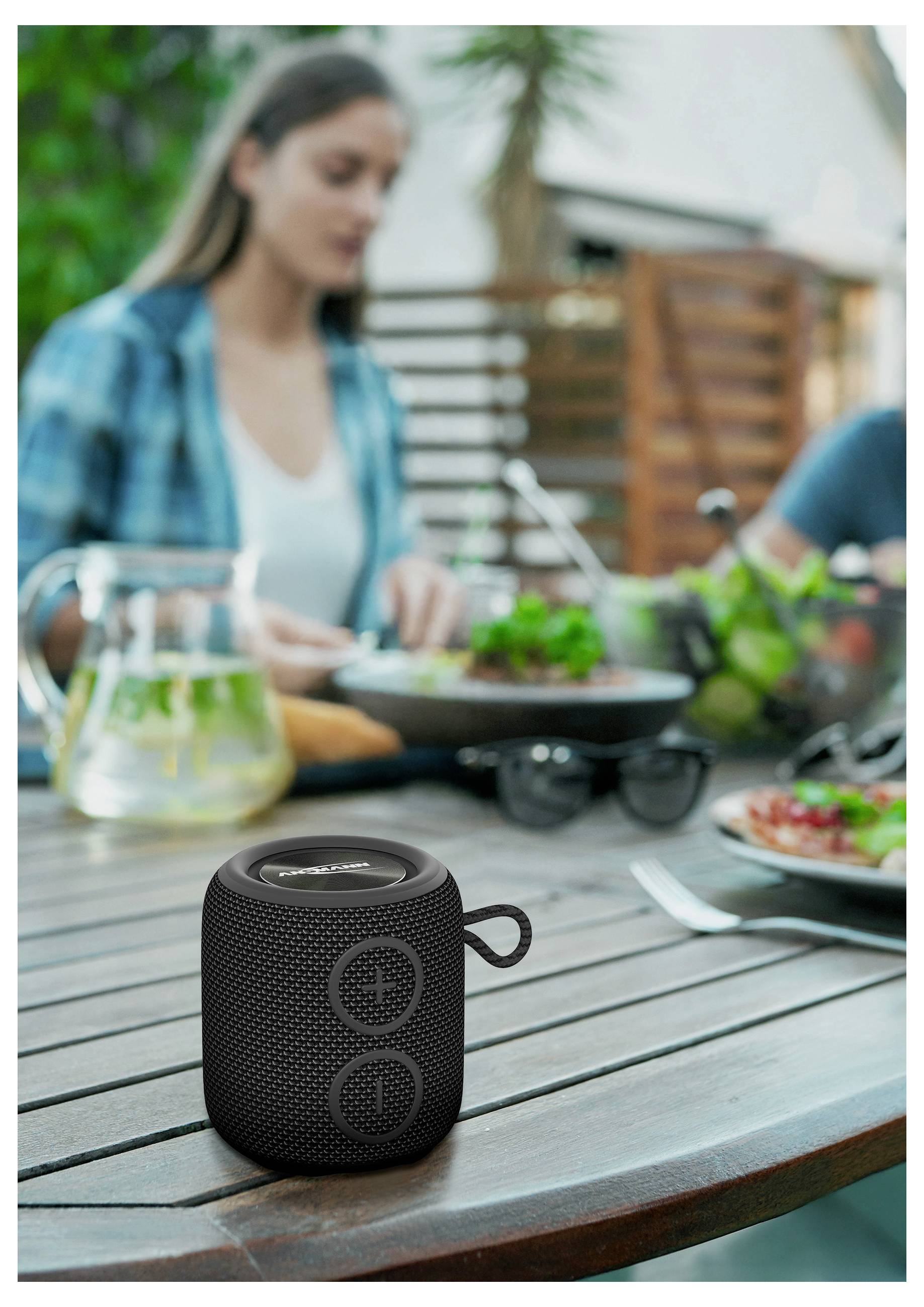 A black portable speaker is on a wooden table outdoors. In the background, a person dining, suggesting a lively, casual setting.