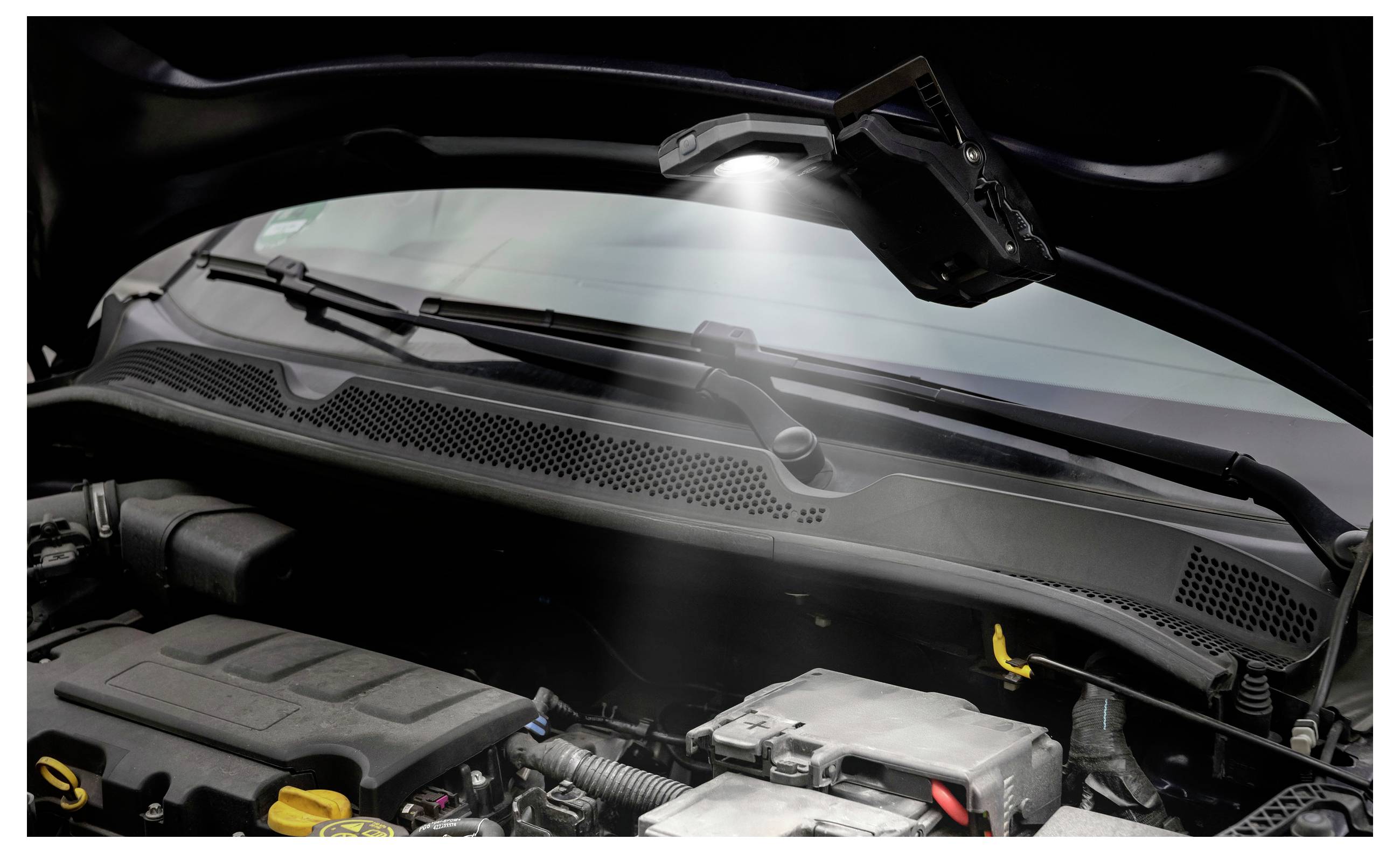 A car engine compartment with the hood open, illuminated under a work light. The engine components are clearly visible.