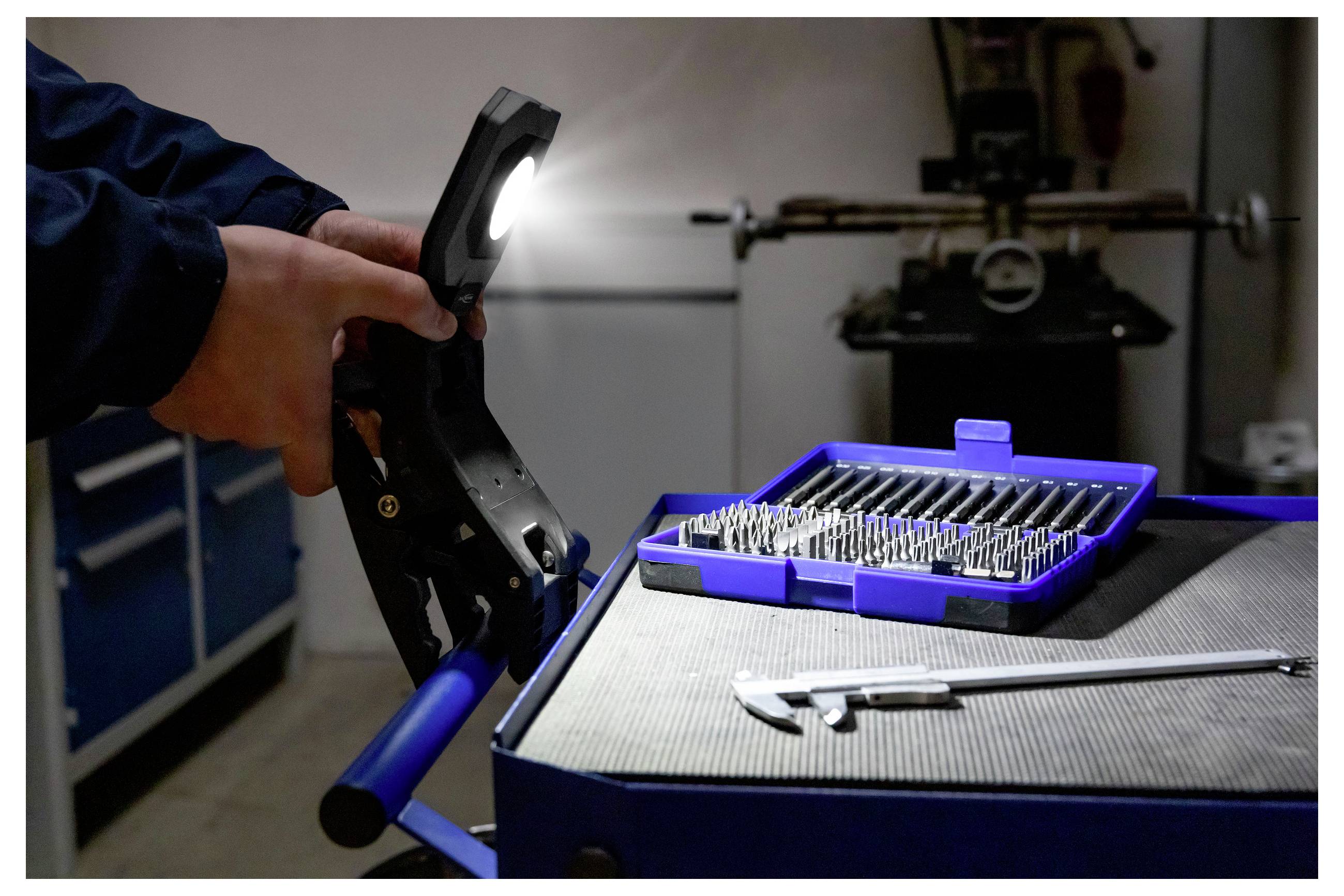 A person holds a tool with a flashlight, illuminating a blue box of drill bits on a workbench next to a caliper in a workshop.