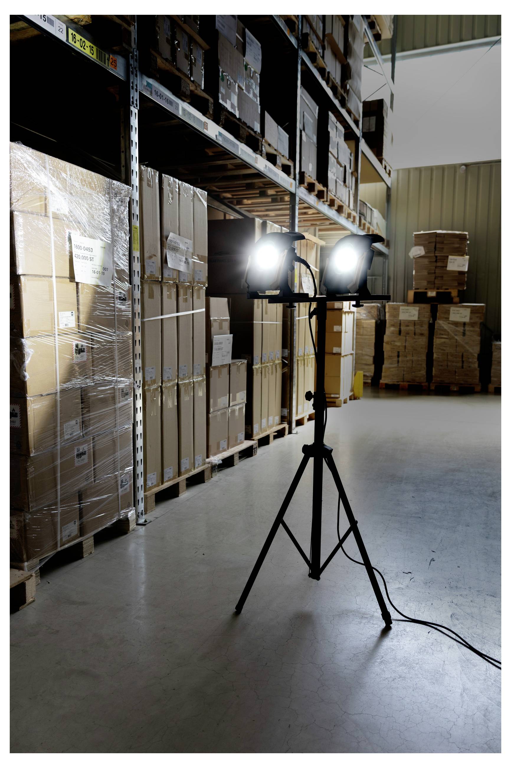 Warehouse interior with shelves stacked with boxes on the left. A tripod with two bright lights illuminates the area in the center.