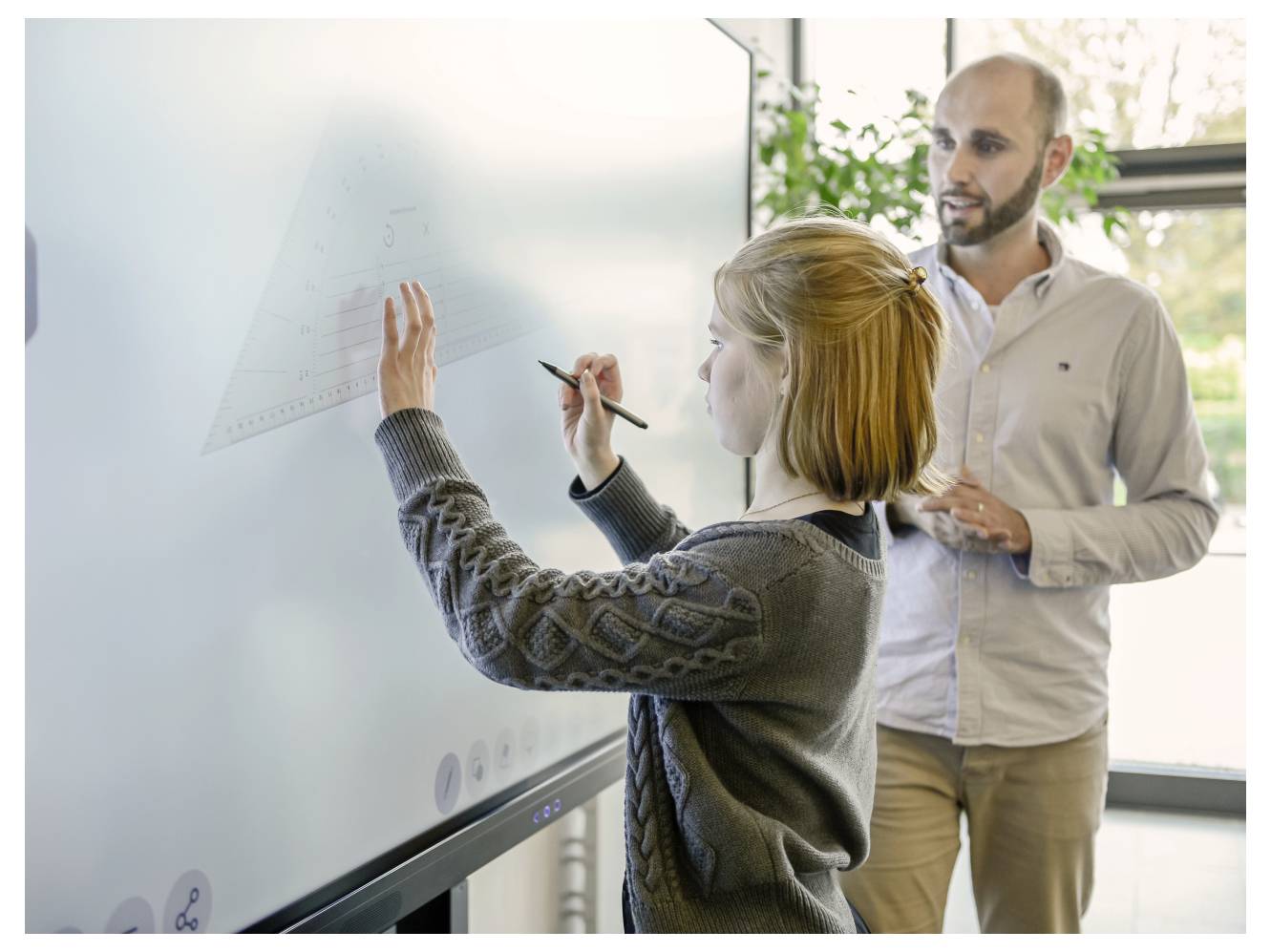 A student writes on an interactive whiteboard with a stylus while a teacher observes, illustrating a tech-assisted learning environment.
