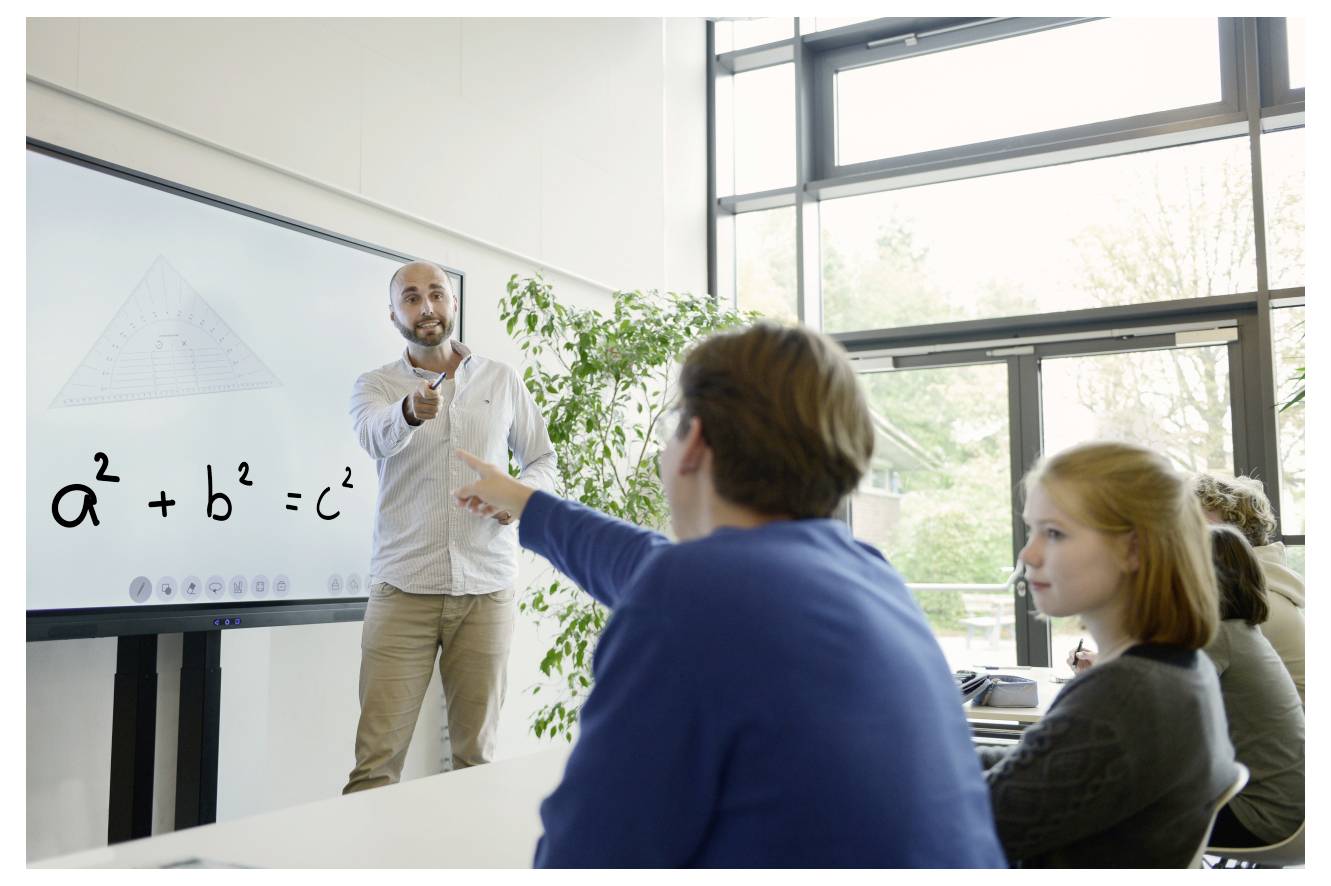 A teacher explains the Pythagorean theorem on a whiteboard to three students in a bright classroom with large windows and potted plants.