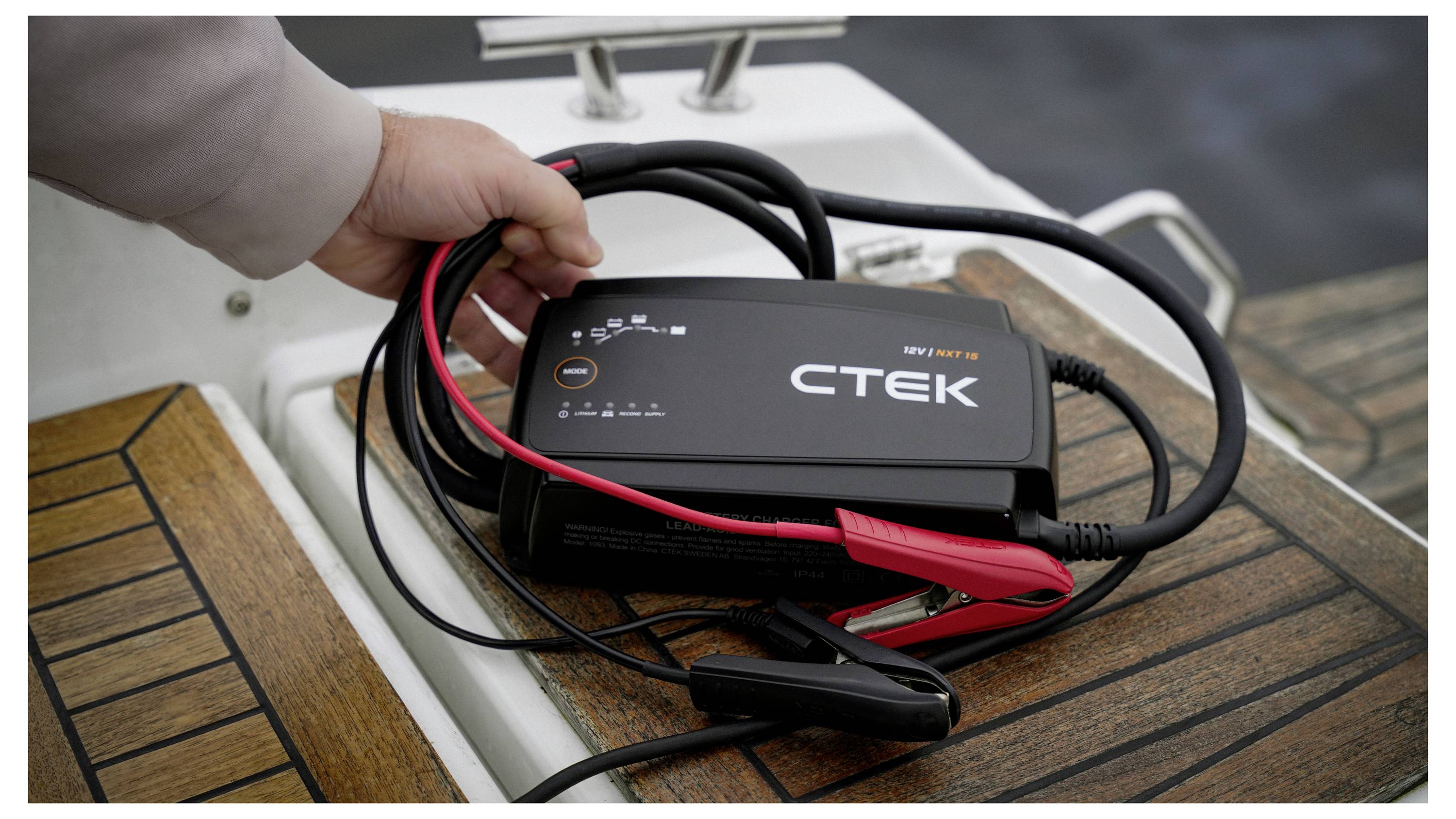 A hand holding a black CTEK battery charger with red and black cables, placed on a boat's wooden deck, suggesting use for marine battery charging.