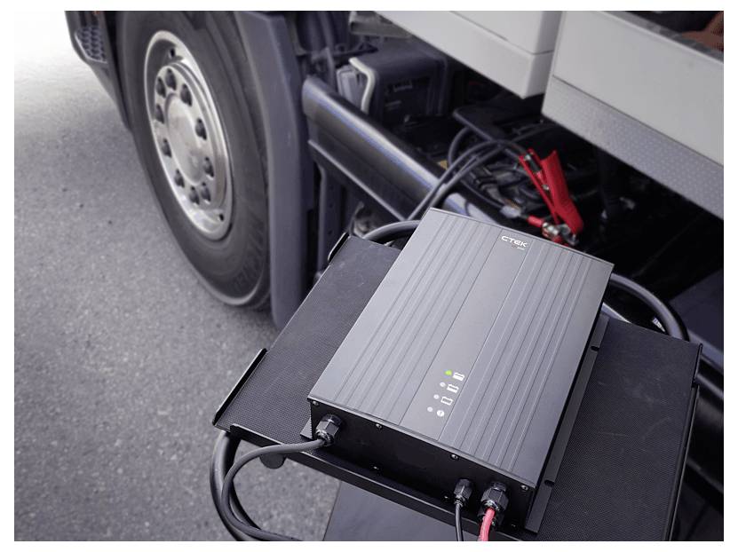 A black charging device with cables connected to a large vehicle. The vehicle's wheel and battery clamp are visible in the background.