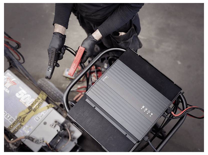 A person with gloves holding jumper cables, preparing to connect a battery charger to a vehicle in a garage setting.