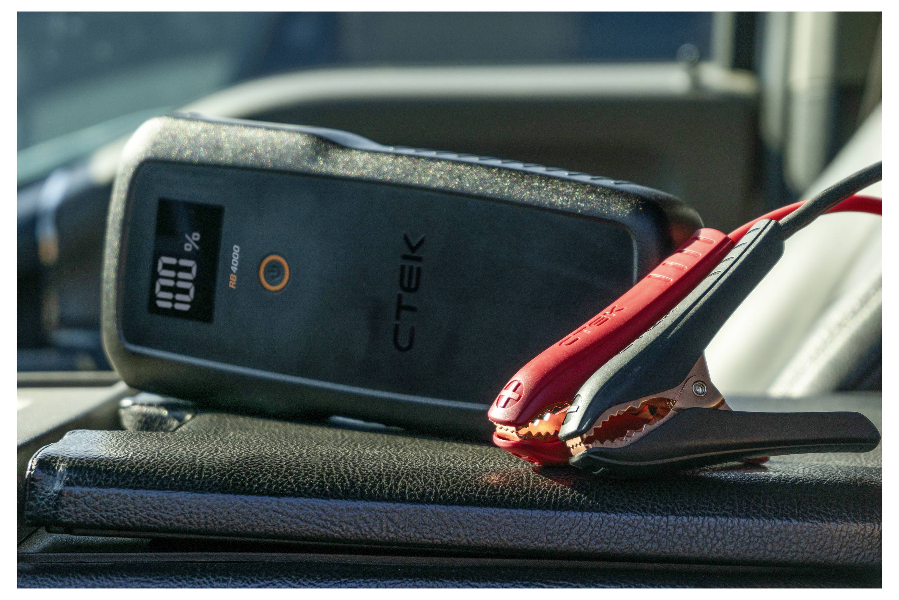 A battery jump starter with red and black clamps is placed on a car dashboard, showing a digital display reading '100%'.
