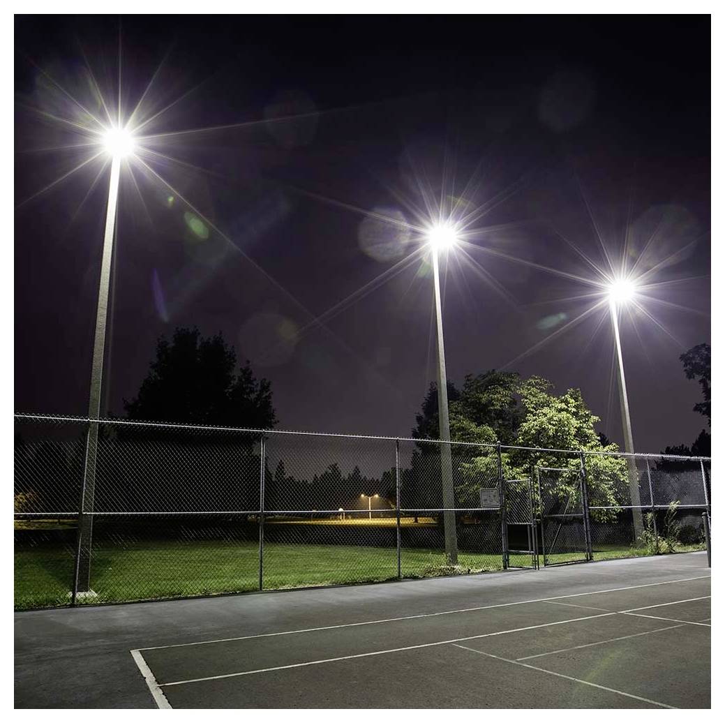 A tennis court at night illuminated by bright overhead lights, surrounded by a chain-link fence, with trees in the background.