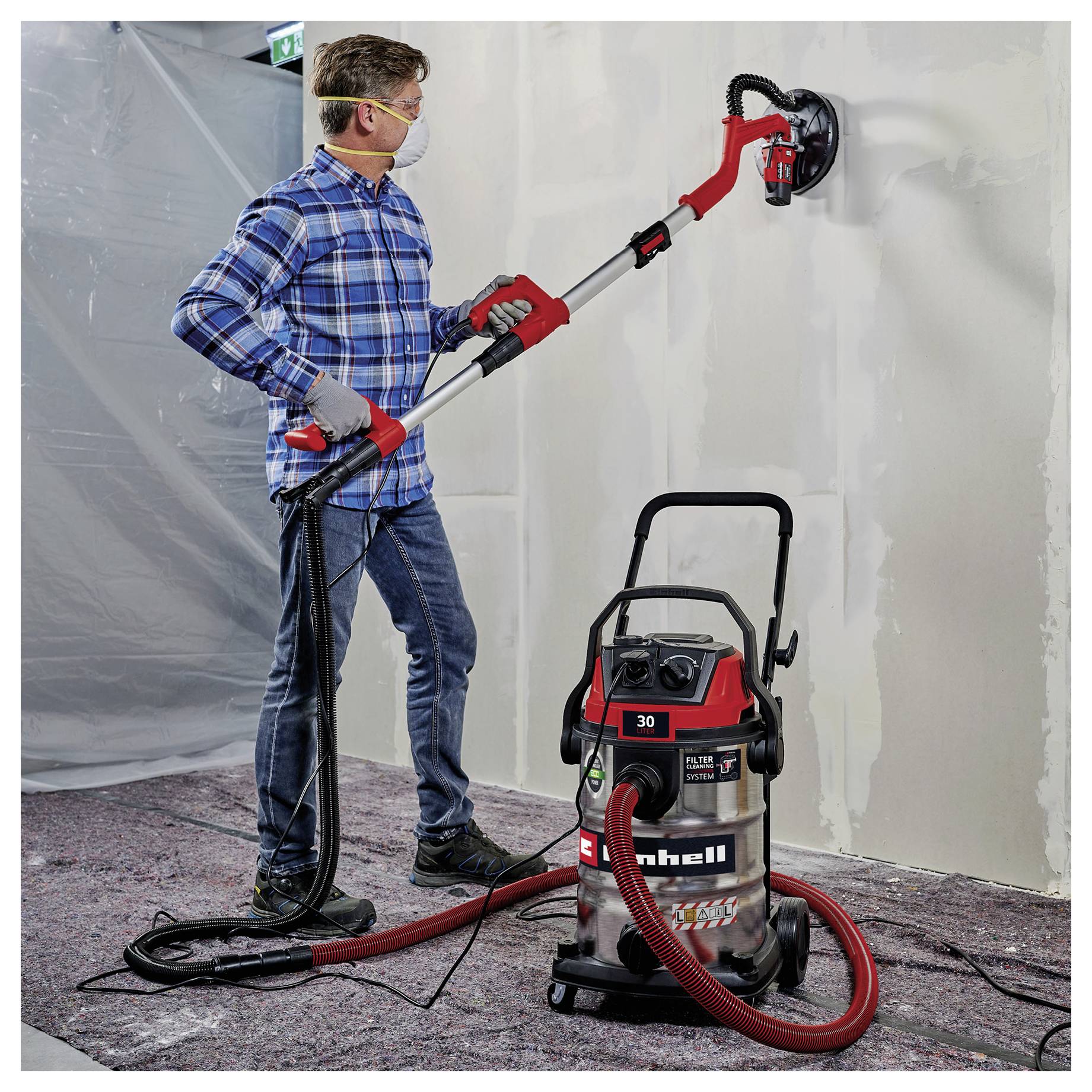 A person wearing a mask uses a red vacuum sander to smooth drywall, with a connected vacuum to collect dust, in an indoor setting.
