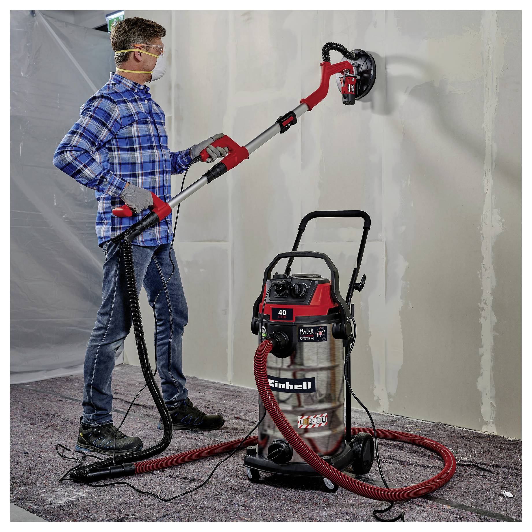 A person wearing safety goggles uses a red and black wall sander connected to a large vacuum, smoothing a drywall surface.