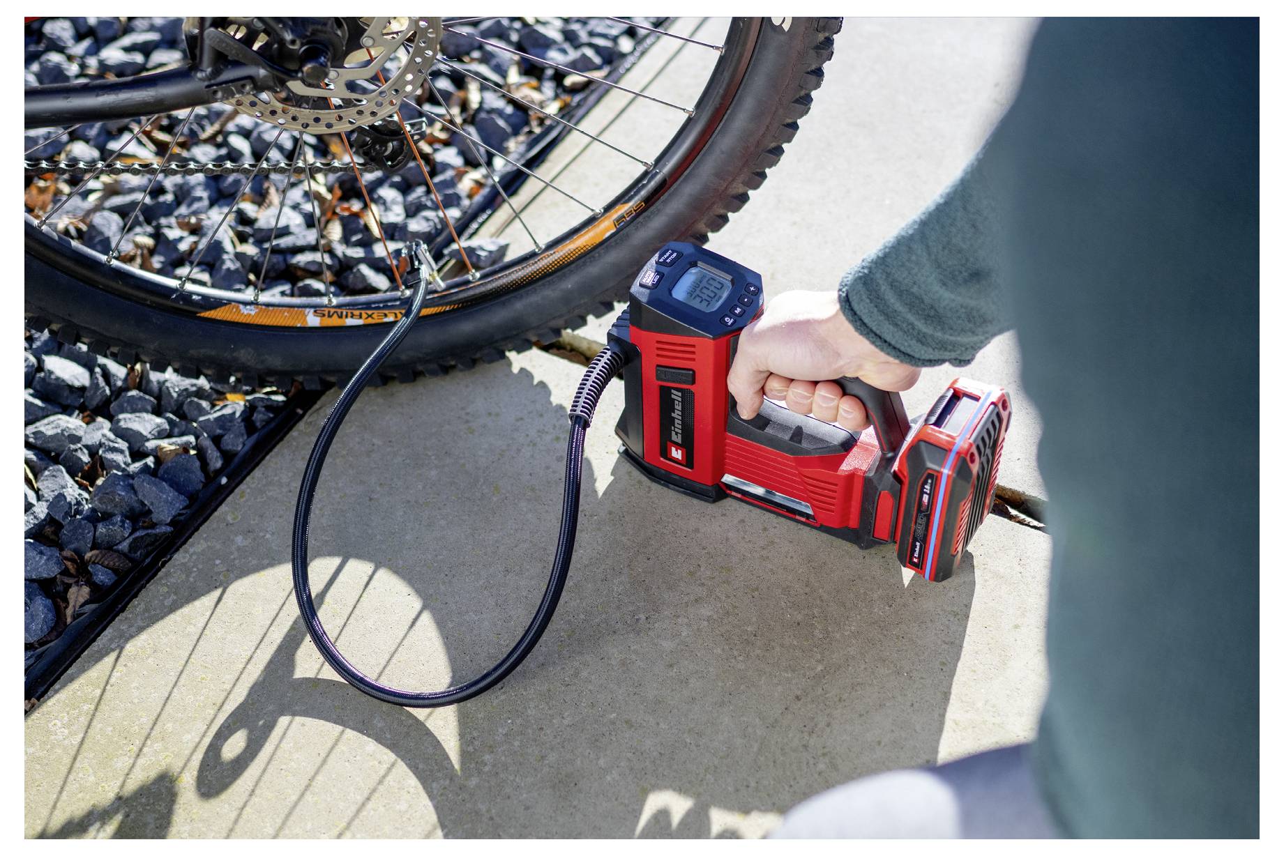 A person uses a red portable air pump to inflate a bicycle tire on a sunny day. The pump's digital display is shown.