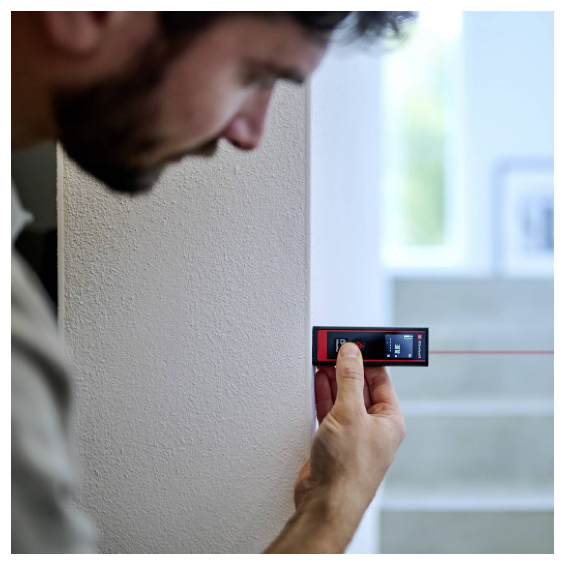 A person uses a digital measuring device against a wall, pointing towards a staircase, indicating measurement taking.