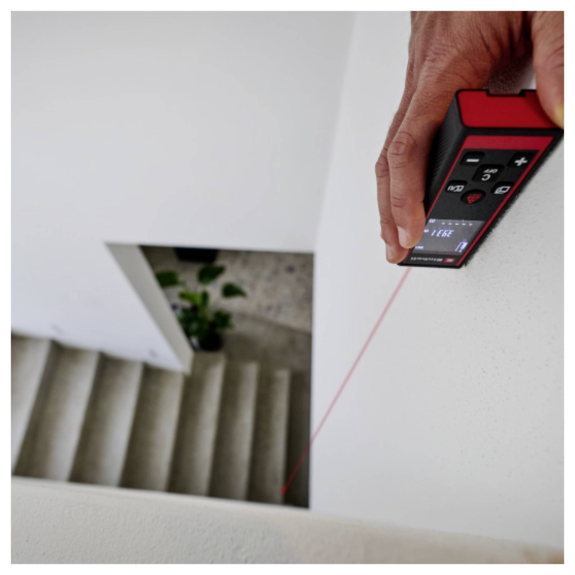 A person uses a red digital laser distance measuring device to measure a vertical drop next to a stairway.